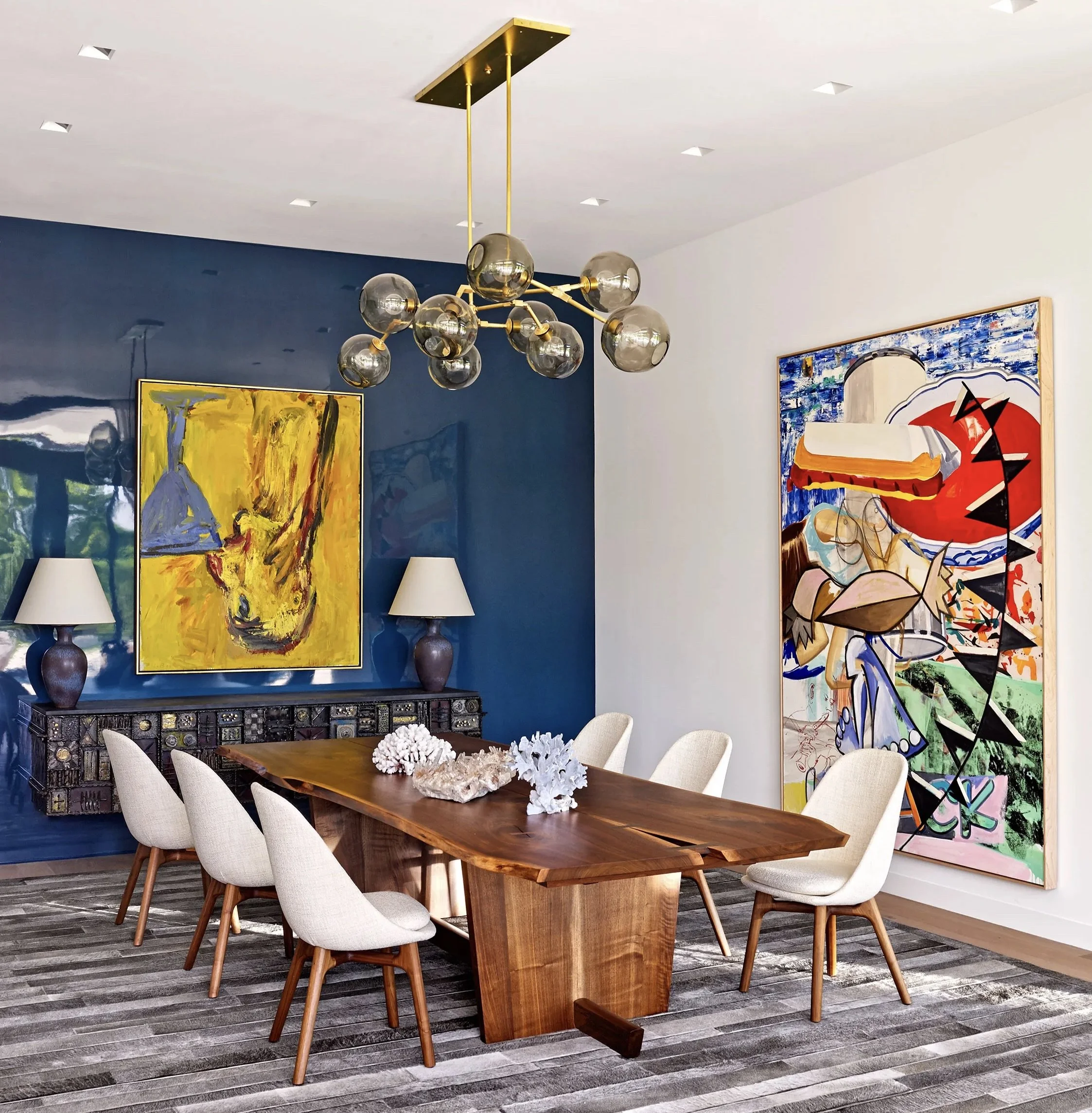 Modern dining room with a wooden table, beige chairs, a dark blue accent wall, abstract paintings, a chandelier with glass globes, and a patterned rug.