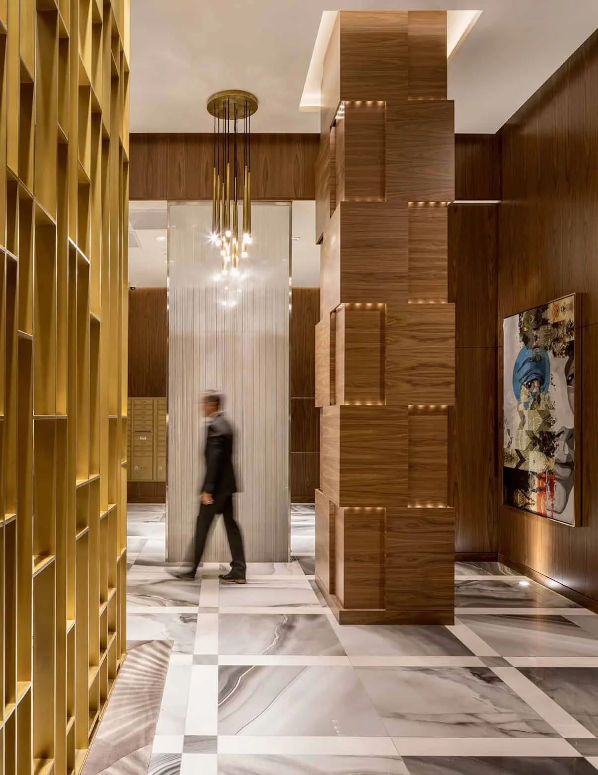 Hotel lobby with marble floors, gold shelving, and wooden walls. A man in a suit is walking past a vertical wooden structure. There is a modern chandelier hanging from the ceiling and a colorful abstract painting on the wall.