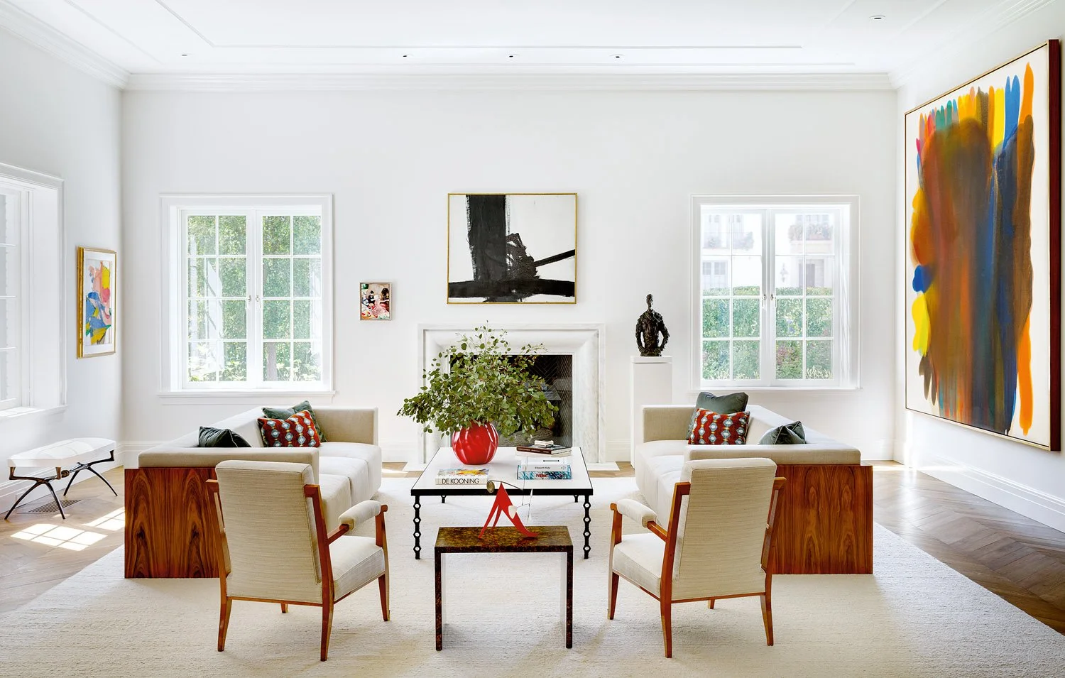 Bright living room with white walls, large windows, leather armchairs, contemporary artwork, a fireplace, and a glass table with books and a red vase with green foliage.