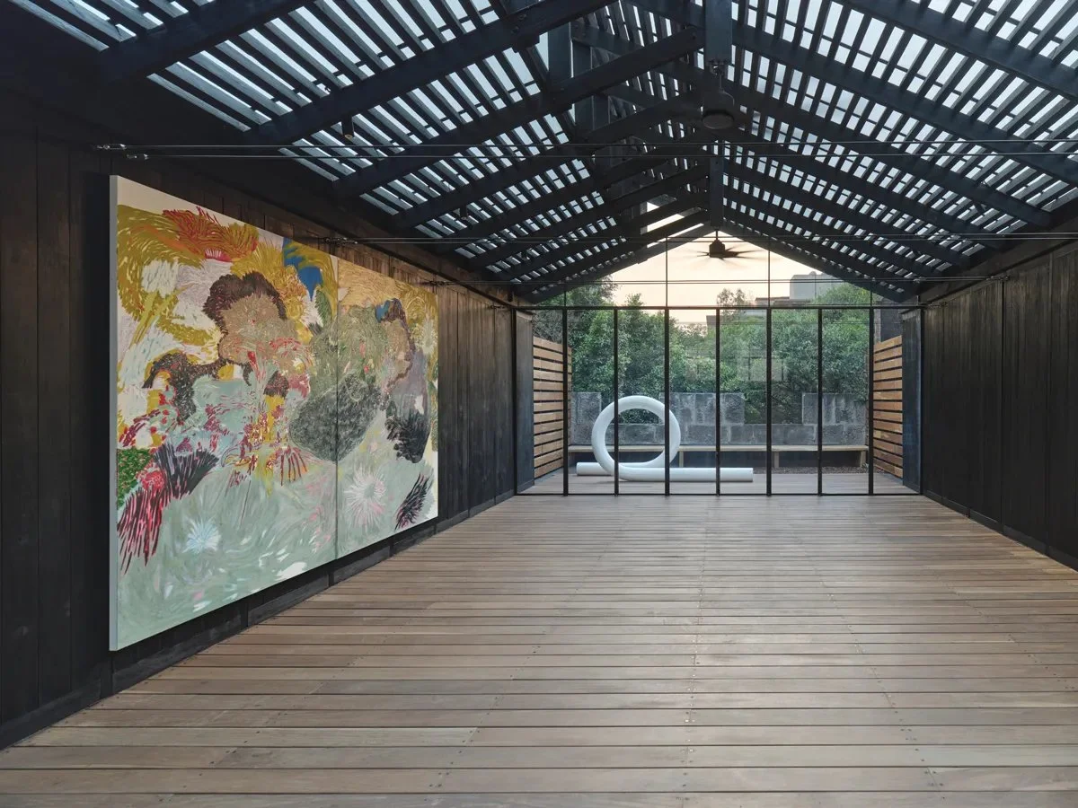 An art gallery with black wooden walls, a wooden floor, a large colorful painting on the left wall, and modern sculptures including a white hollow ring at the far end near glass windows showing trees outside.