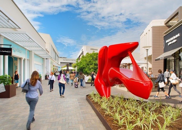 Large red abstract sculpture along an outdoor retail promenade, surrounded by architecture and pedestrians.