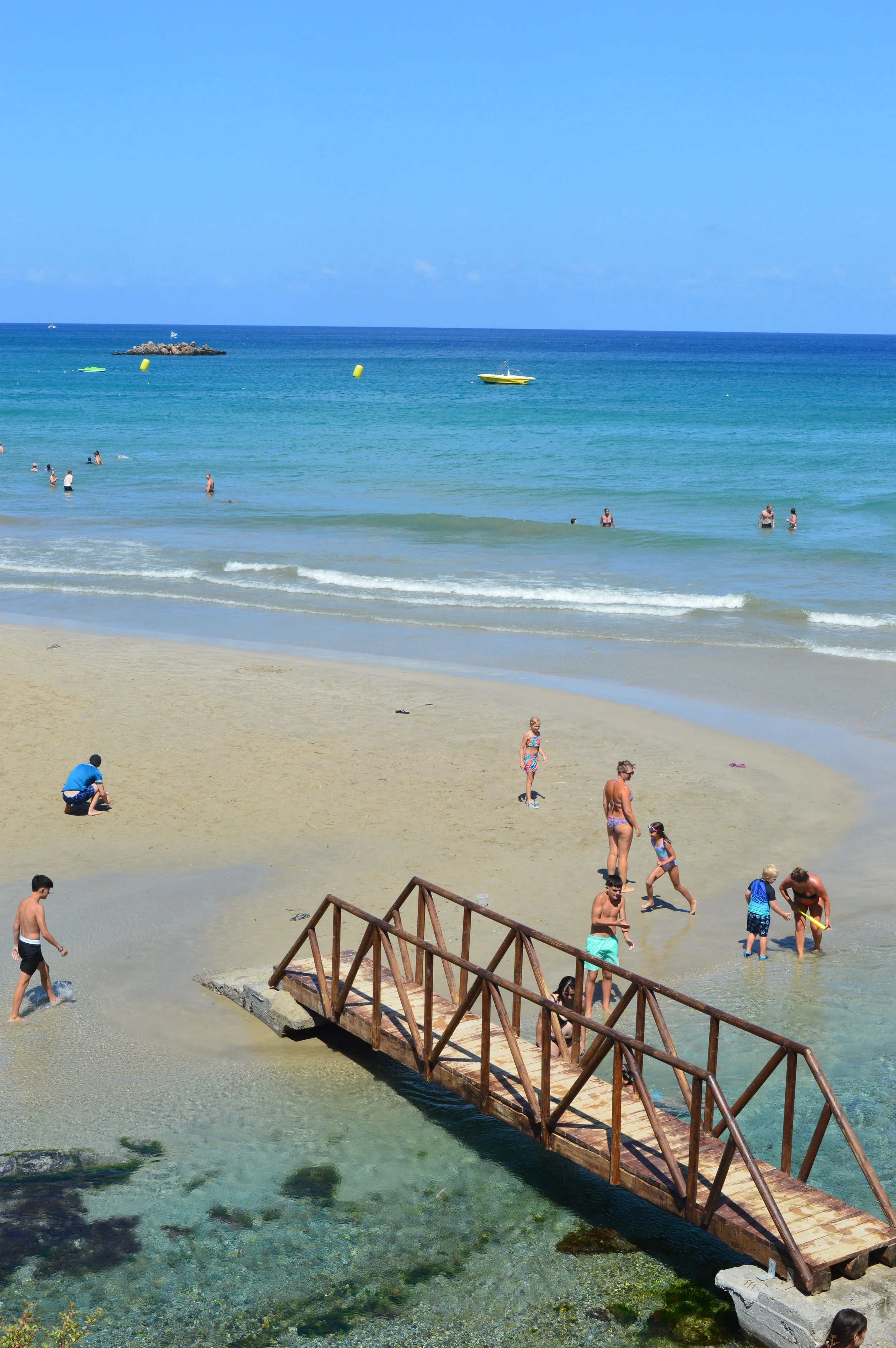 People enjoy the beach, some swimming in the ocean, others walking on the sand, with a small wooden bridge over a shallow area of water in the foreground and boats floating in the distance.