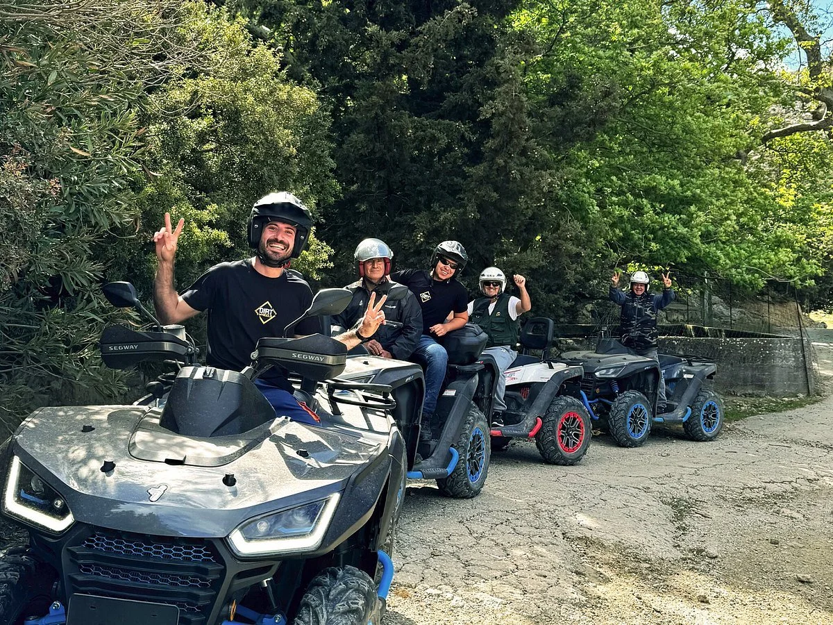 A group of five people wearing helmets and riding all-terrain vehicles (ATVs) on a dirt path surrounded by green trees, smiling and making peace signs.