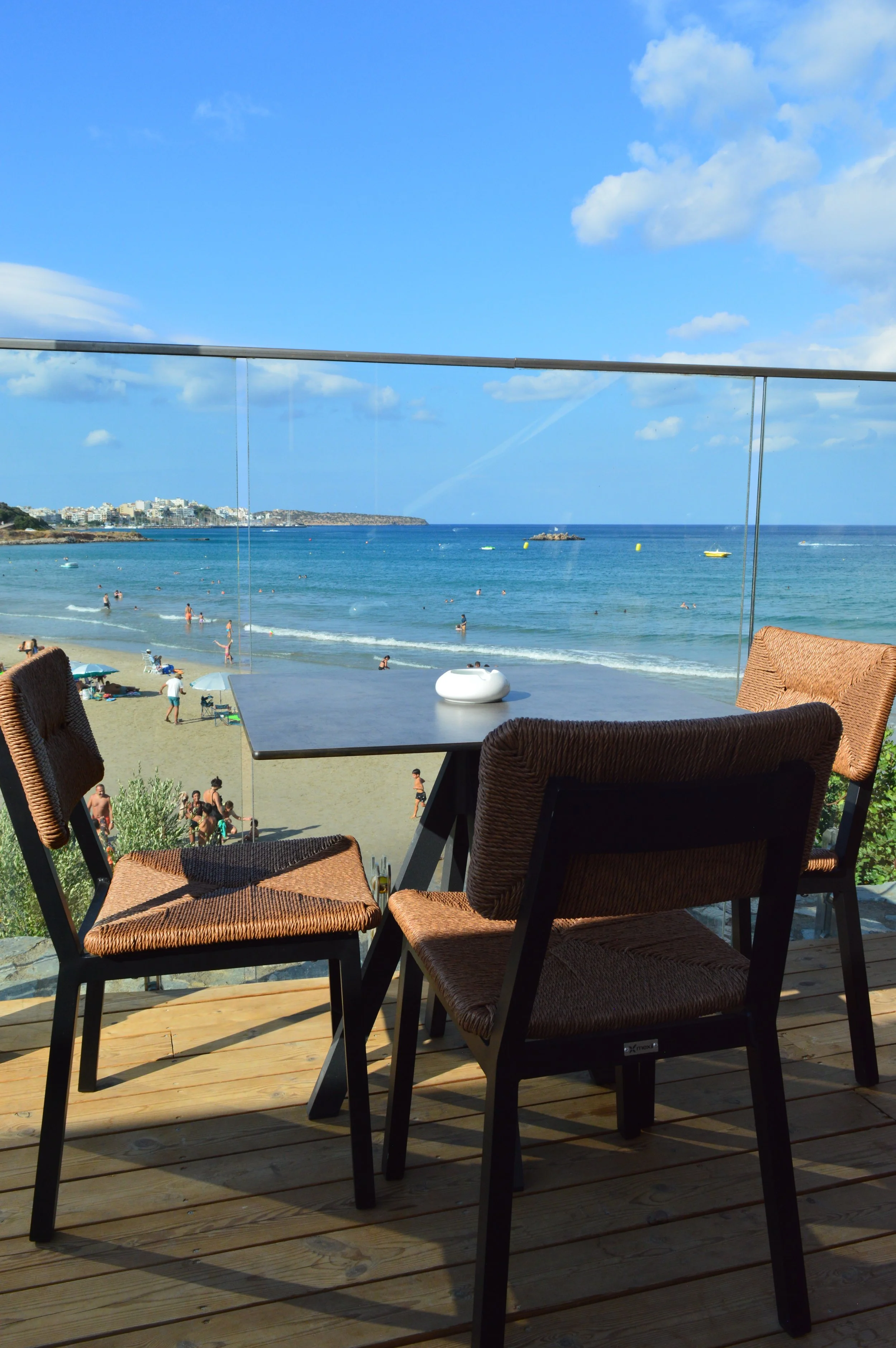A balcony with three wicker chairs and a black table overlooking a busy beach and ocean under a partly cloudy blue sky.