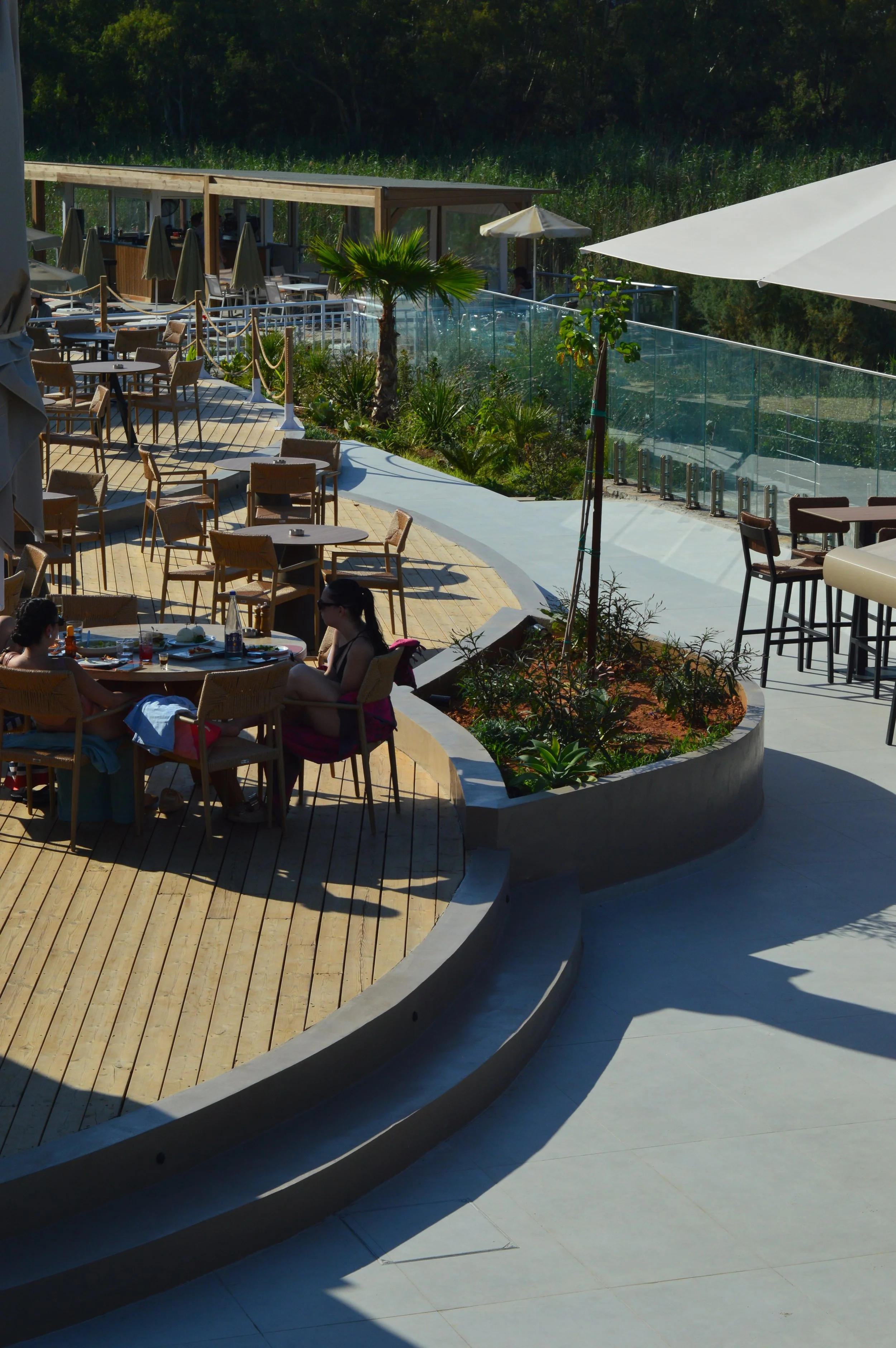 Outdoor restaurant patio with wooden flooring, tables, chairs, umbrellas, and some people dining, surrounded by greenery and trees.