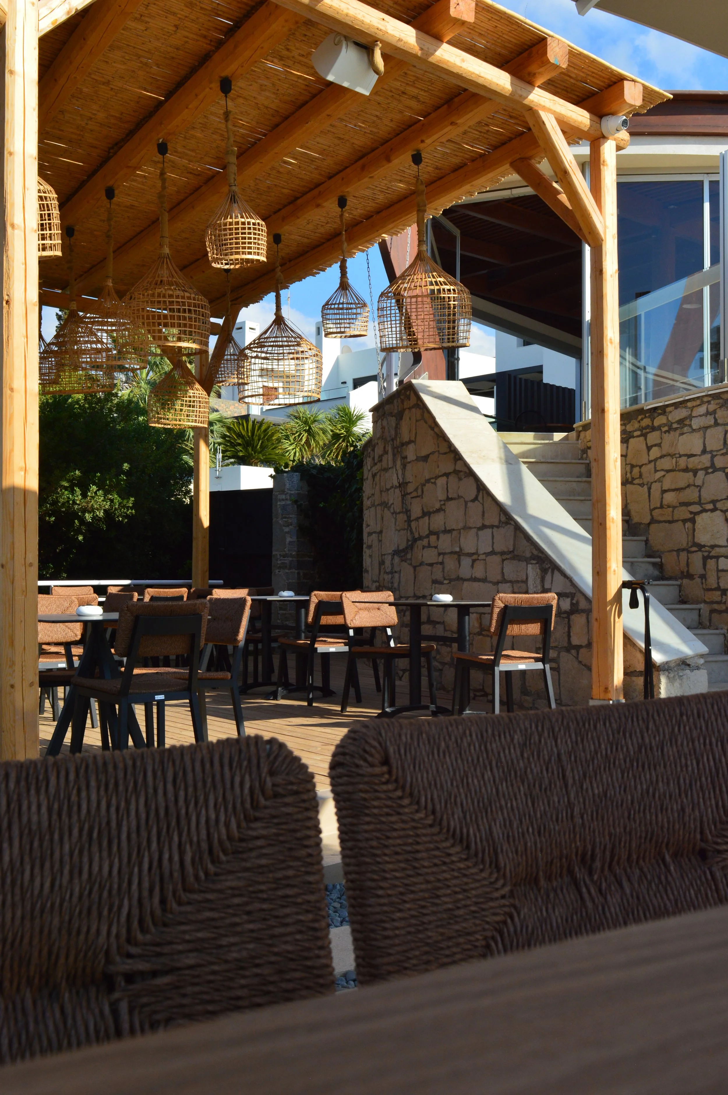 Outdoor patio with woven chair backs in the foreground, wooden tables and chairs under a wooden pergola, hanging wicker lanterns, stone staircase, and green foliage in the background.