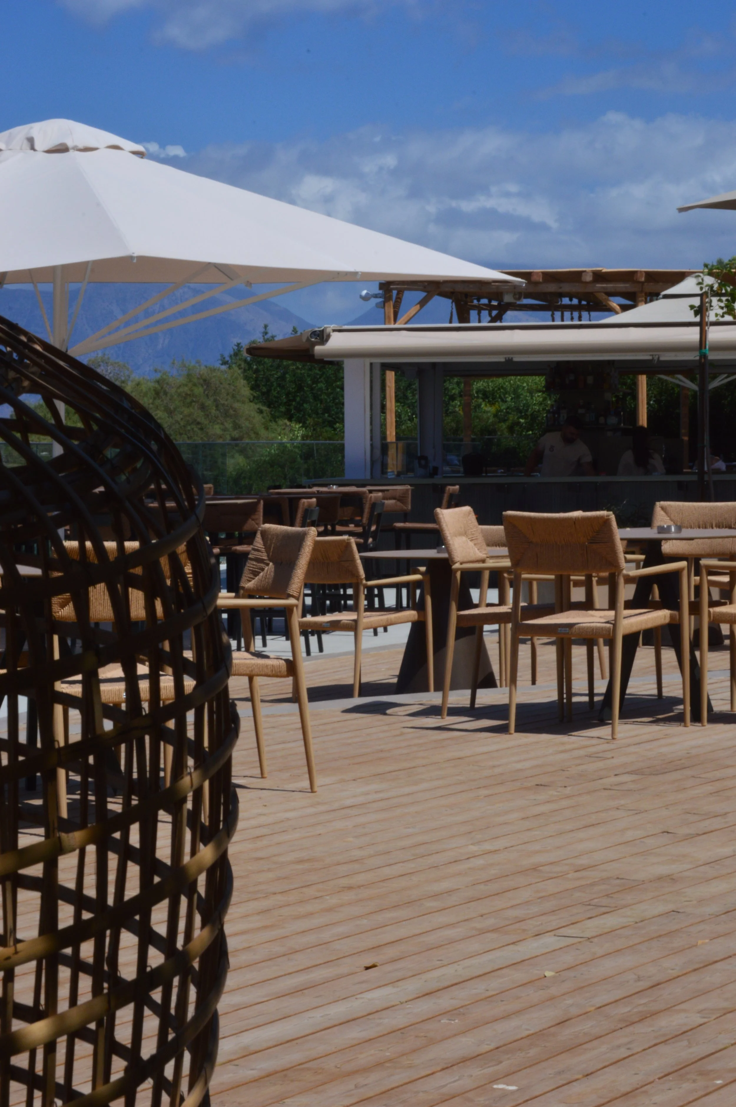 Empty outdoor restaurant patio with wooden tables and chairs, large white umbrellas, and mountains in the distance under a partly cloudy sky.