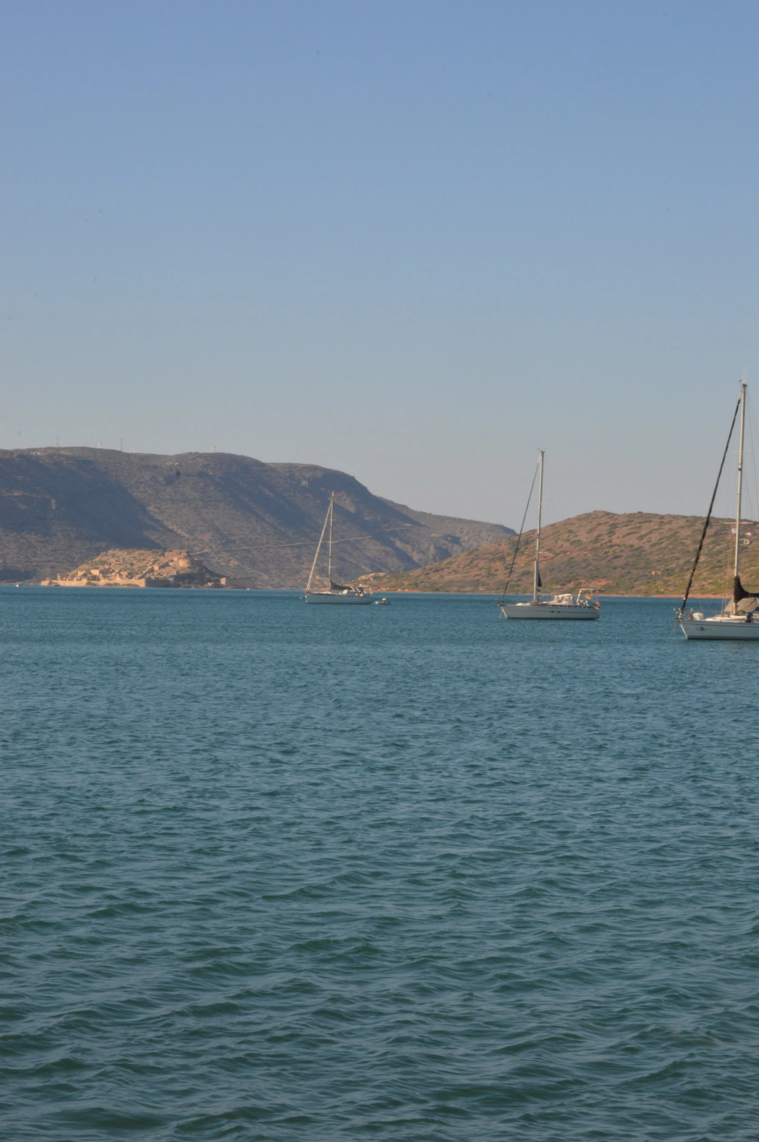 Calm ocean with sailboats anchored near hilly coastline under clear blue sky.