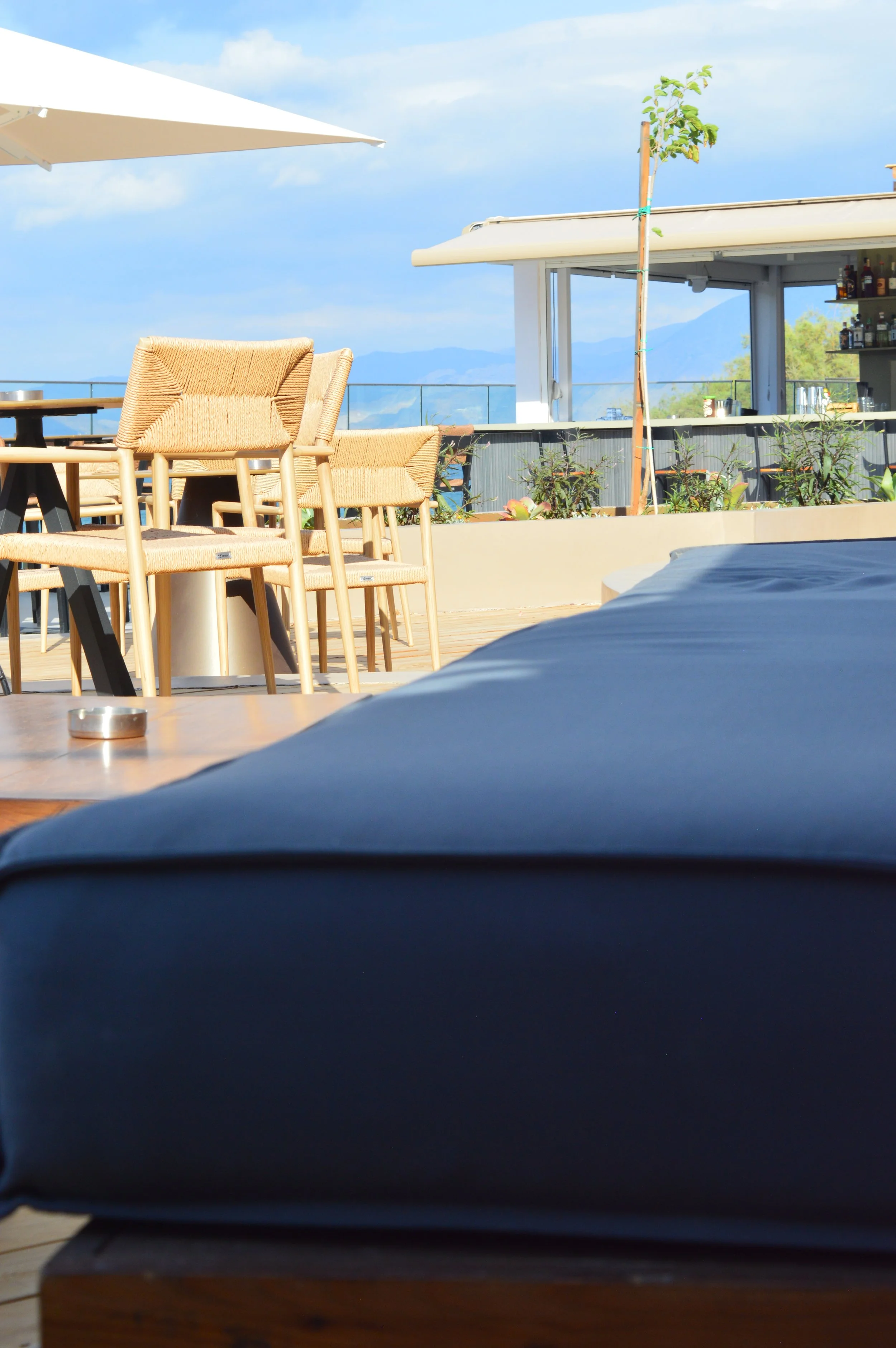 Outdoor rooftop bar or lounge area with wicker chairs, tables, a large umbrella, potted plants, and a bar in the background, with mountains and a blue sky with clouds in the distance.