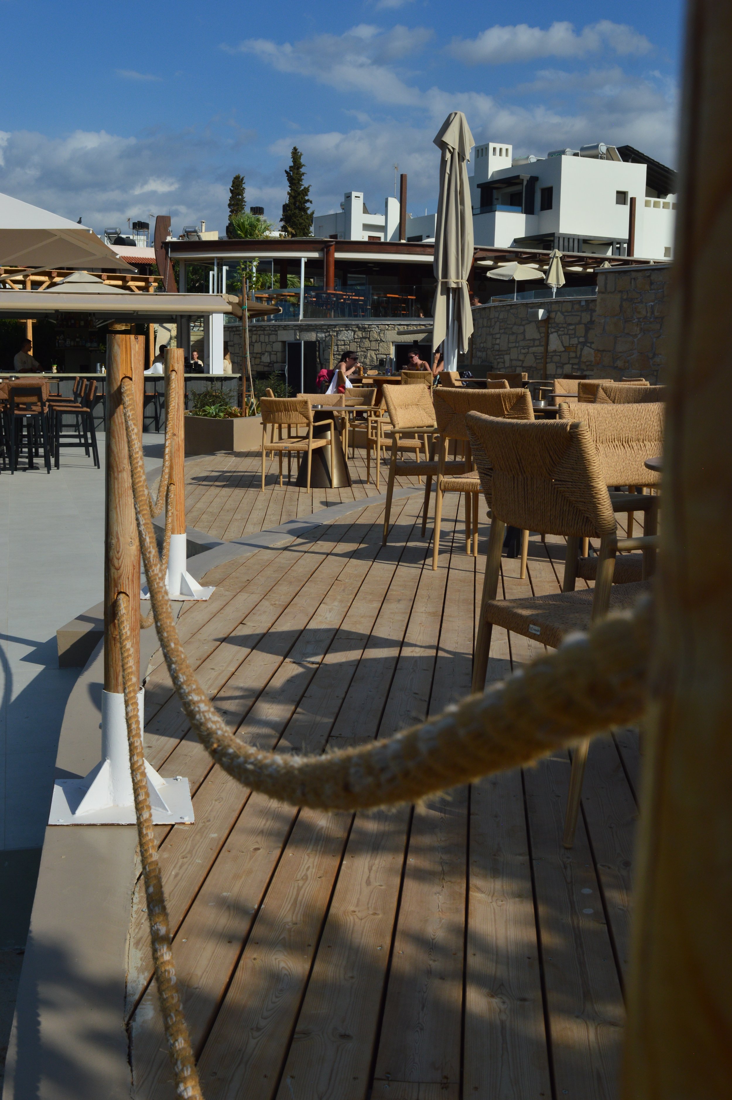An outdoor restaurant or café patio with wooden flooring, beige chairs, and tables. Some patrons are seated, and large umbrellas are visible. In the background, there are modern white buildings and a partly cloudy blue sky.