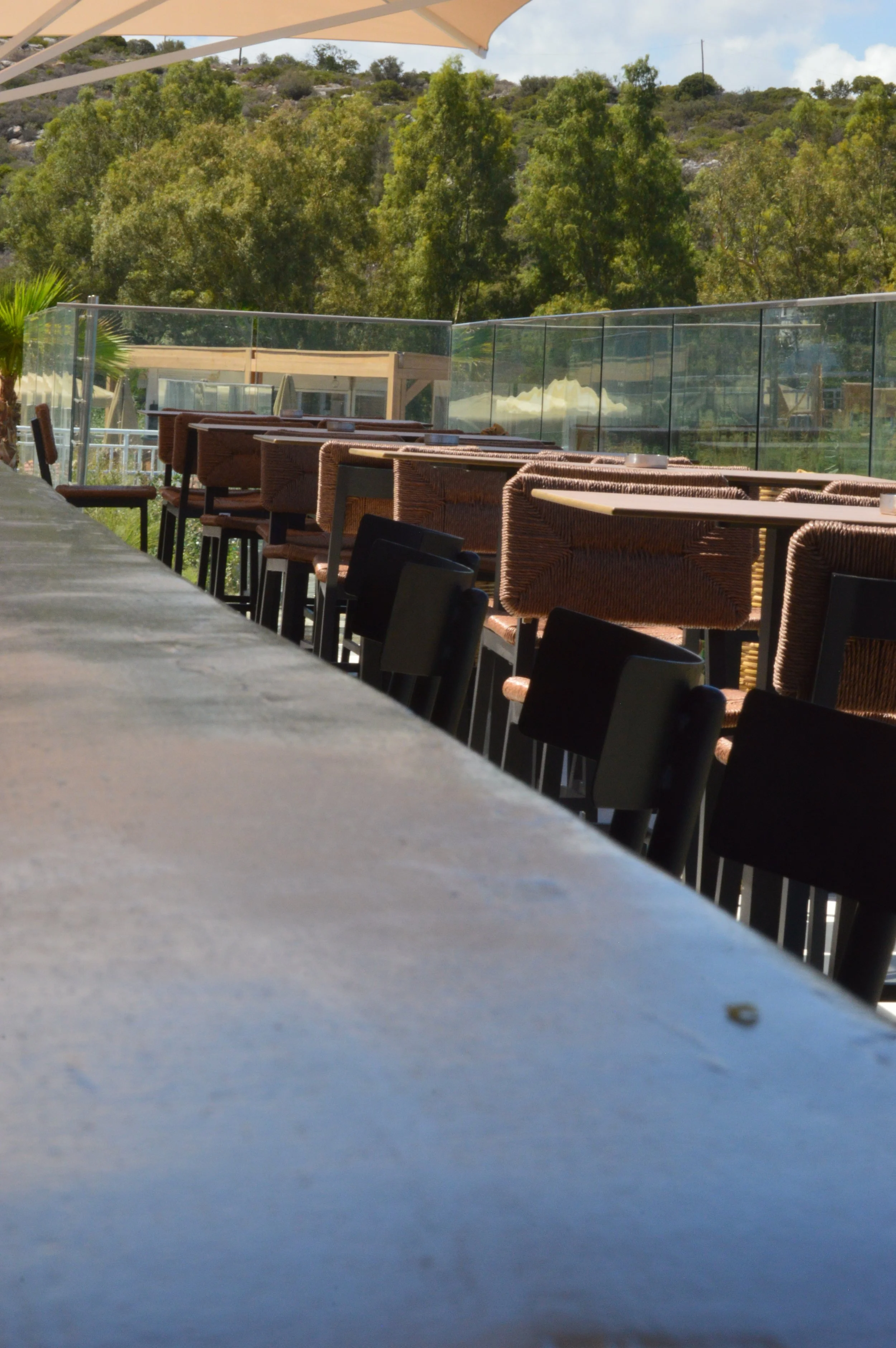Outdoor seating area at a restaurant or cafe with tables, chairs, and a glass railing, overlooking a hillside with trees and shrubs.