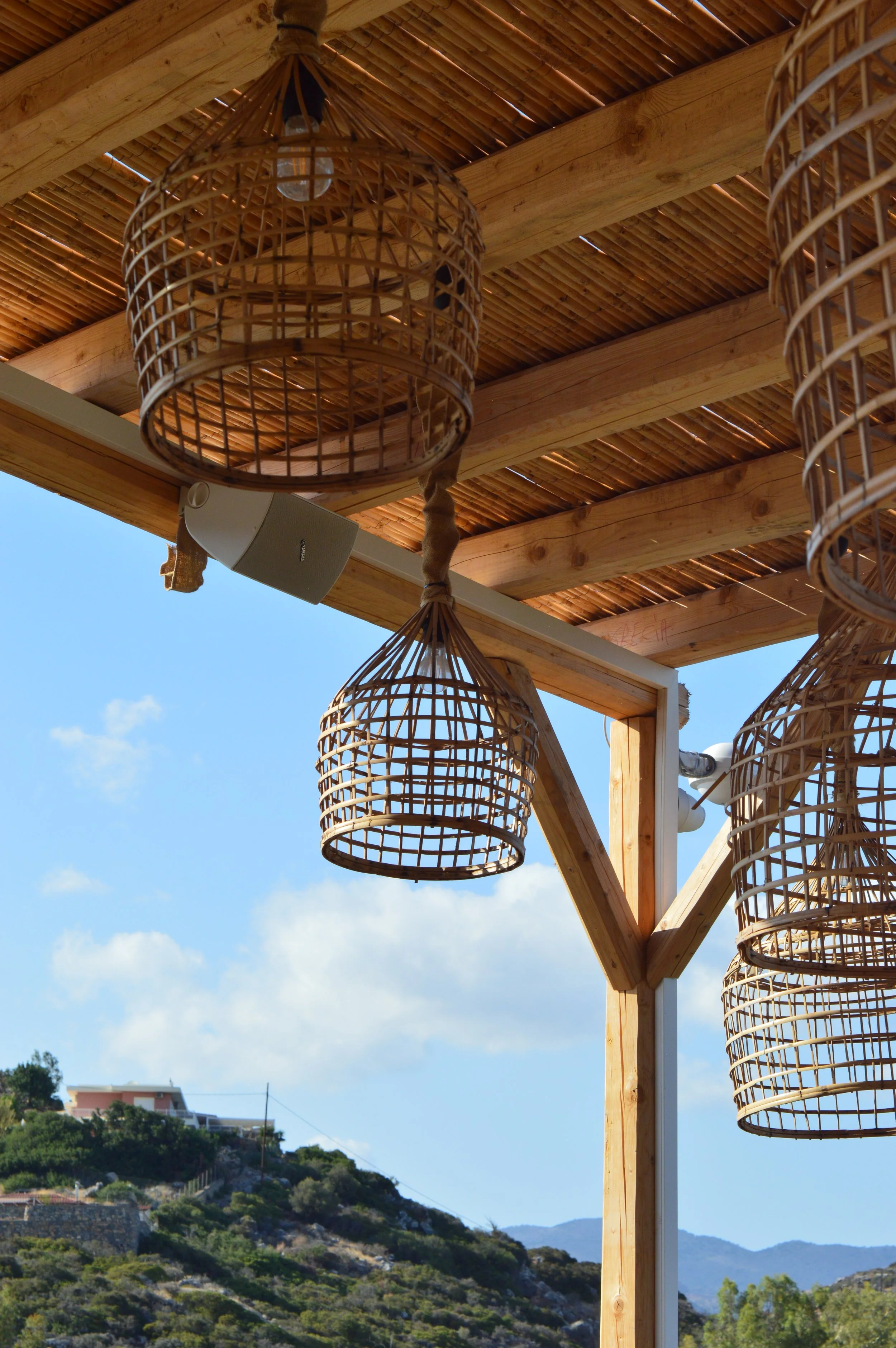 Wicker pendant lights hanging from a wooden ceiling with a clear blue sky and hillside in the background.
