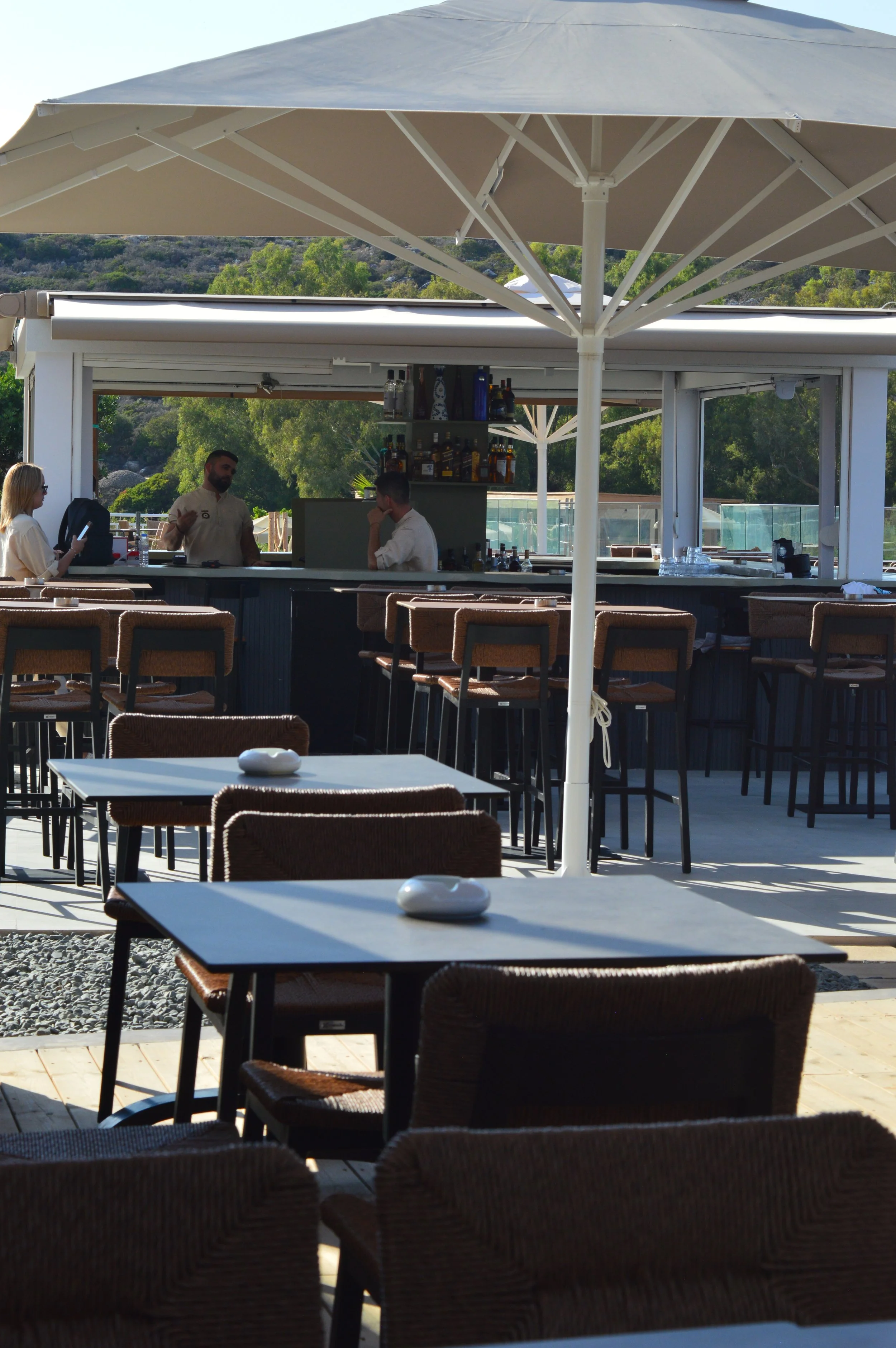 An outdoor bar area with a large white patio umbrella, brown chairs, and tables with white ashtrays, overlooking a landscape with green trees and hills.