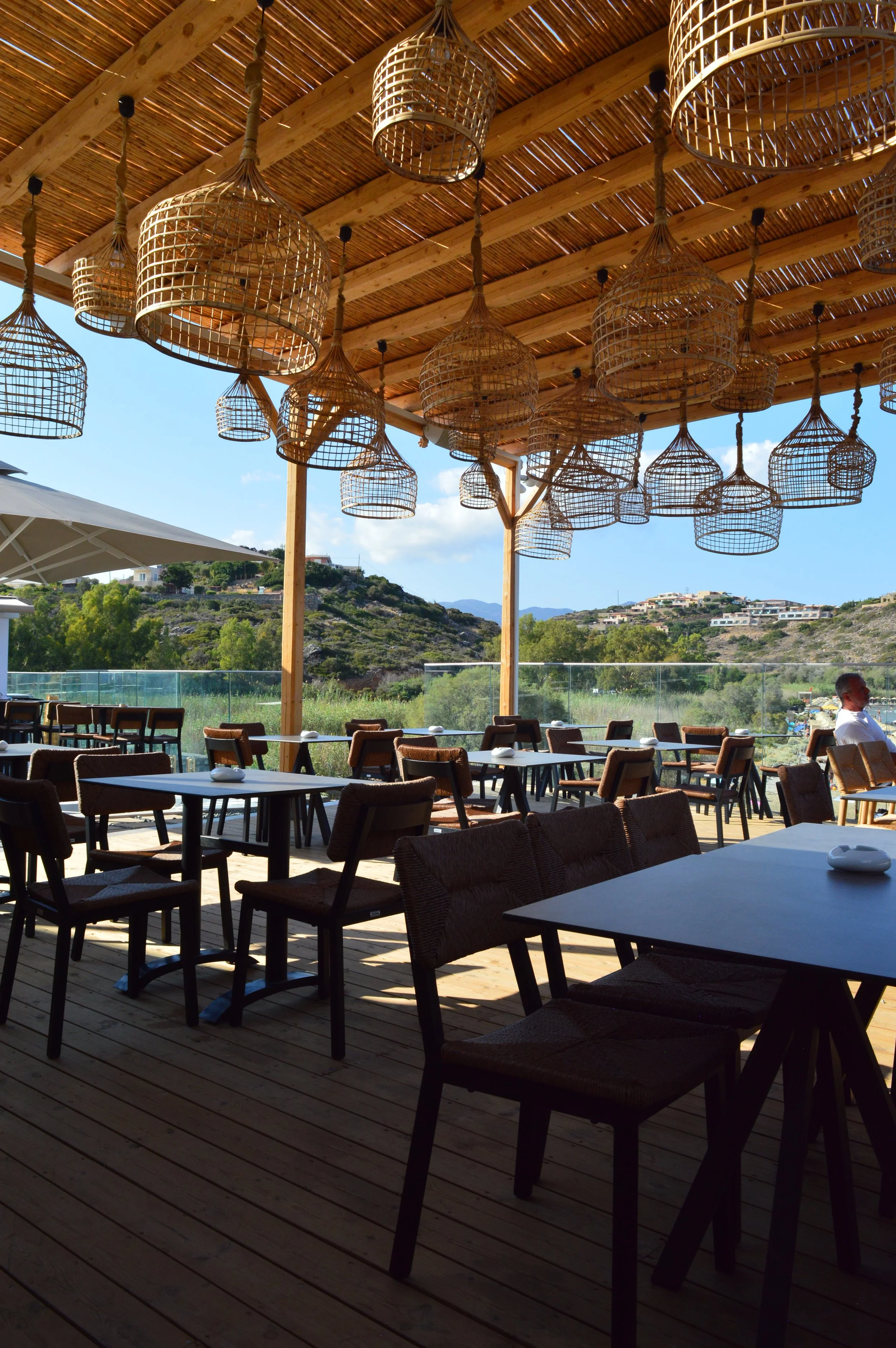 Outdoor restaurant with wooden ceiling, hanging wicker pendant lights, tables, chairs, and scenic hillside view.