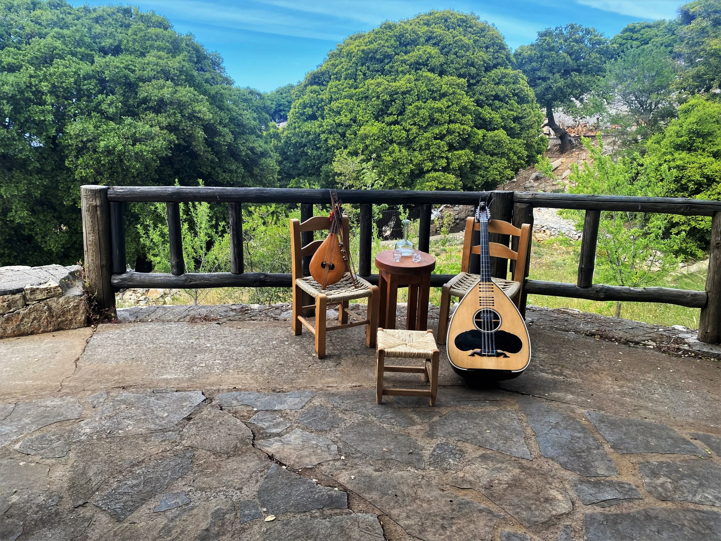 Two wooden chairs with woven seats, a small round wooden table with two glasses and a bottle, and two stringed musical instruments on the ground, set on a stone patio with a wooden railing and a lush green forest in the background.