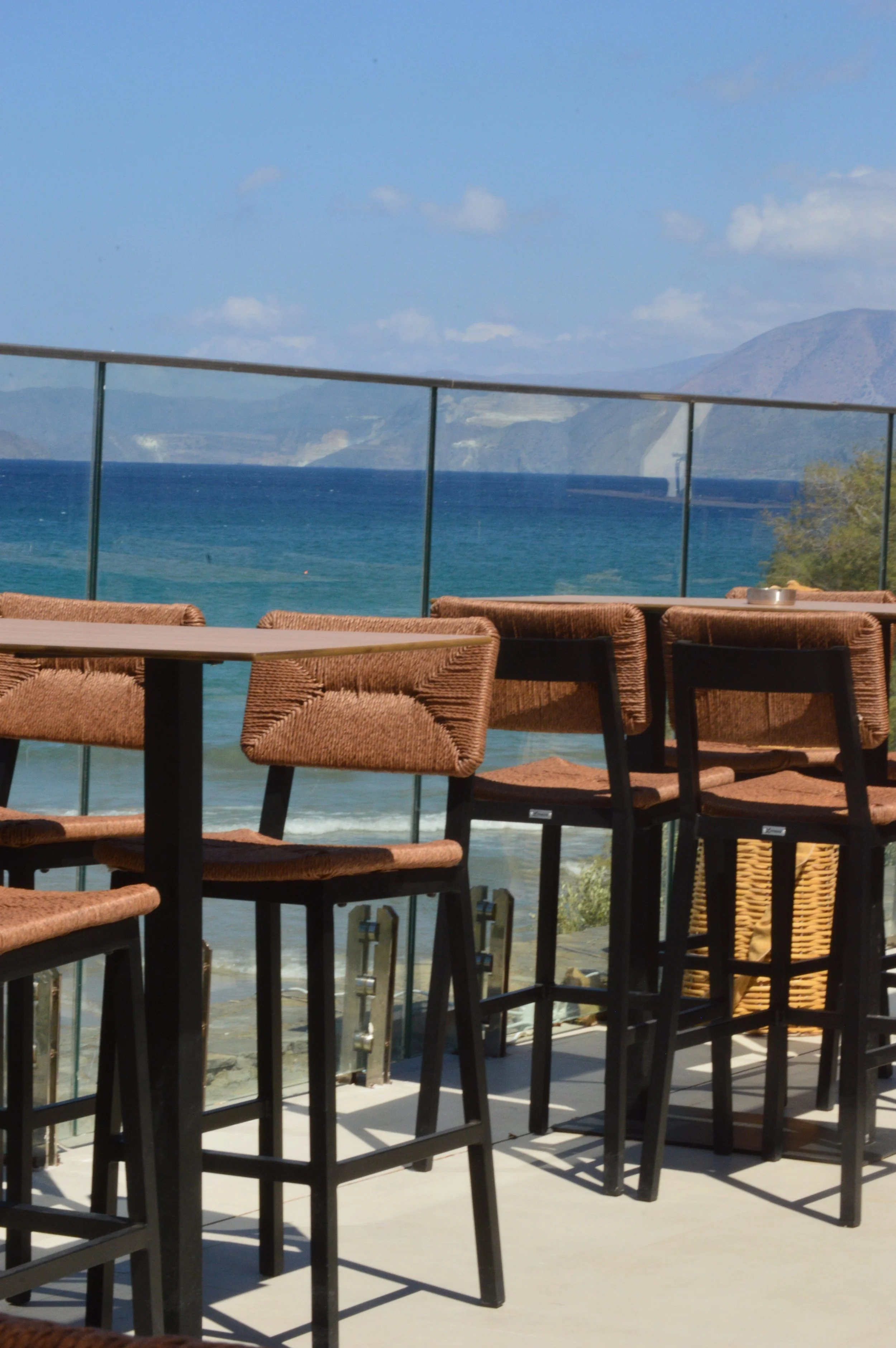 Outdoor seating area with bar stools and tables overlooking the ocean and distant mountains under a blue sky.