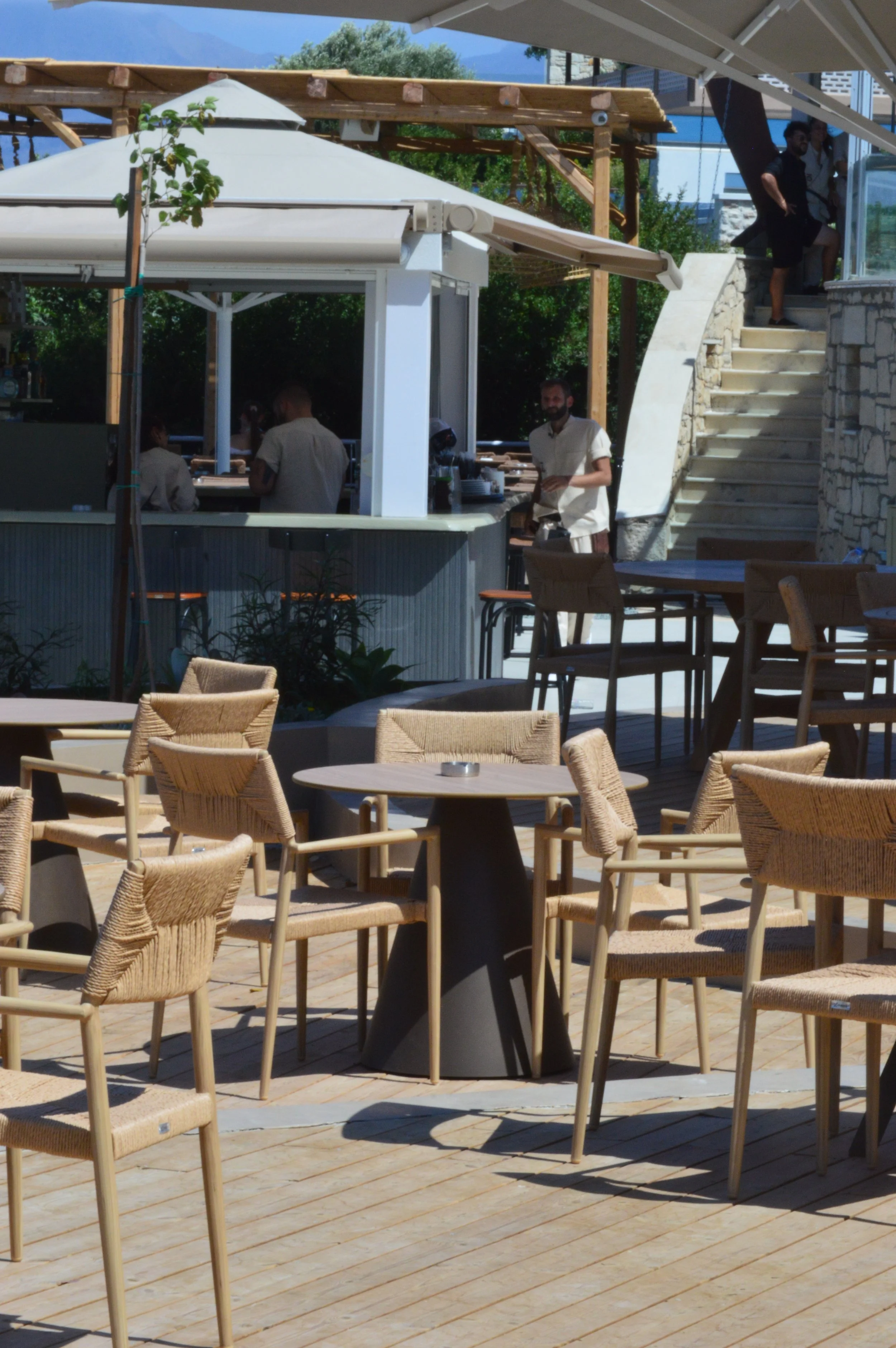 Outdoor seating area at a restaurant with wooden chairs and tables, a covered bar area, and a staircase leading to an upper level, on a sunny day.