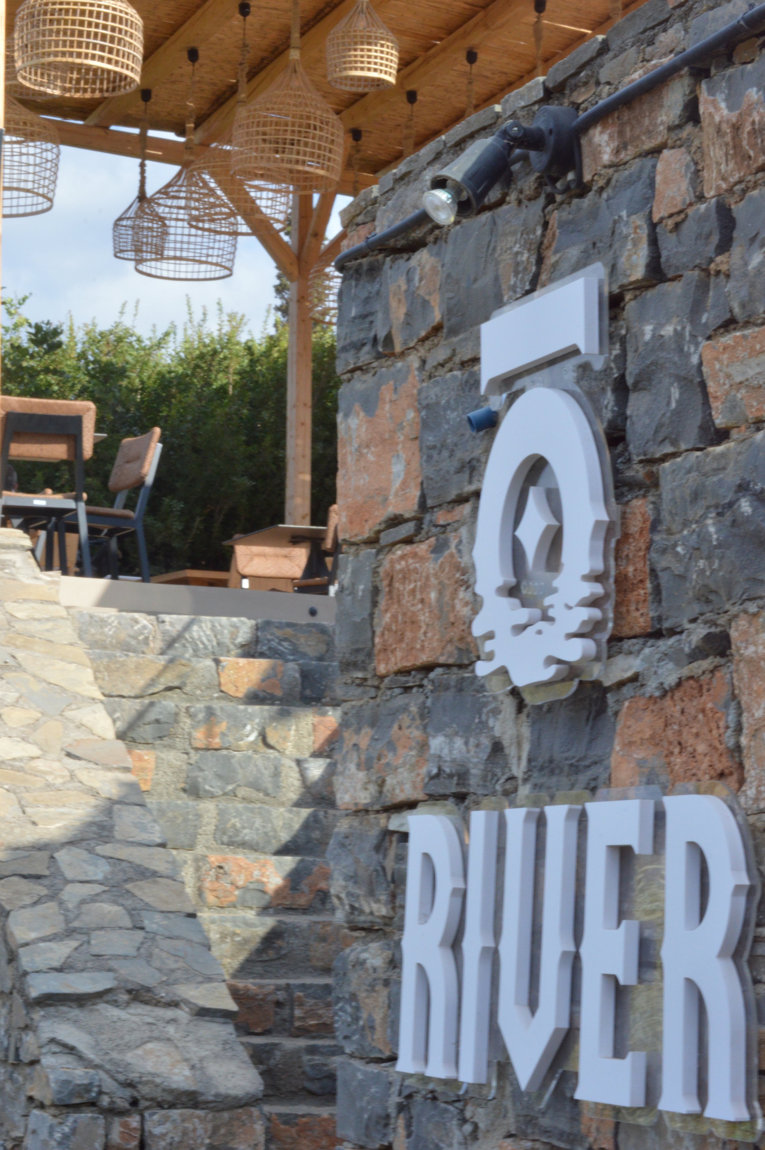 A stone wall with a decorative sign that reads 'RIVER' and a stylized wave design. Behind it, there are outdoor chairs and tables, and a wooden pergola with hanging wicker light fixtures.