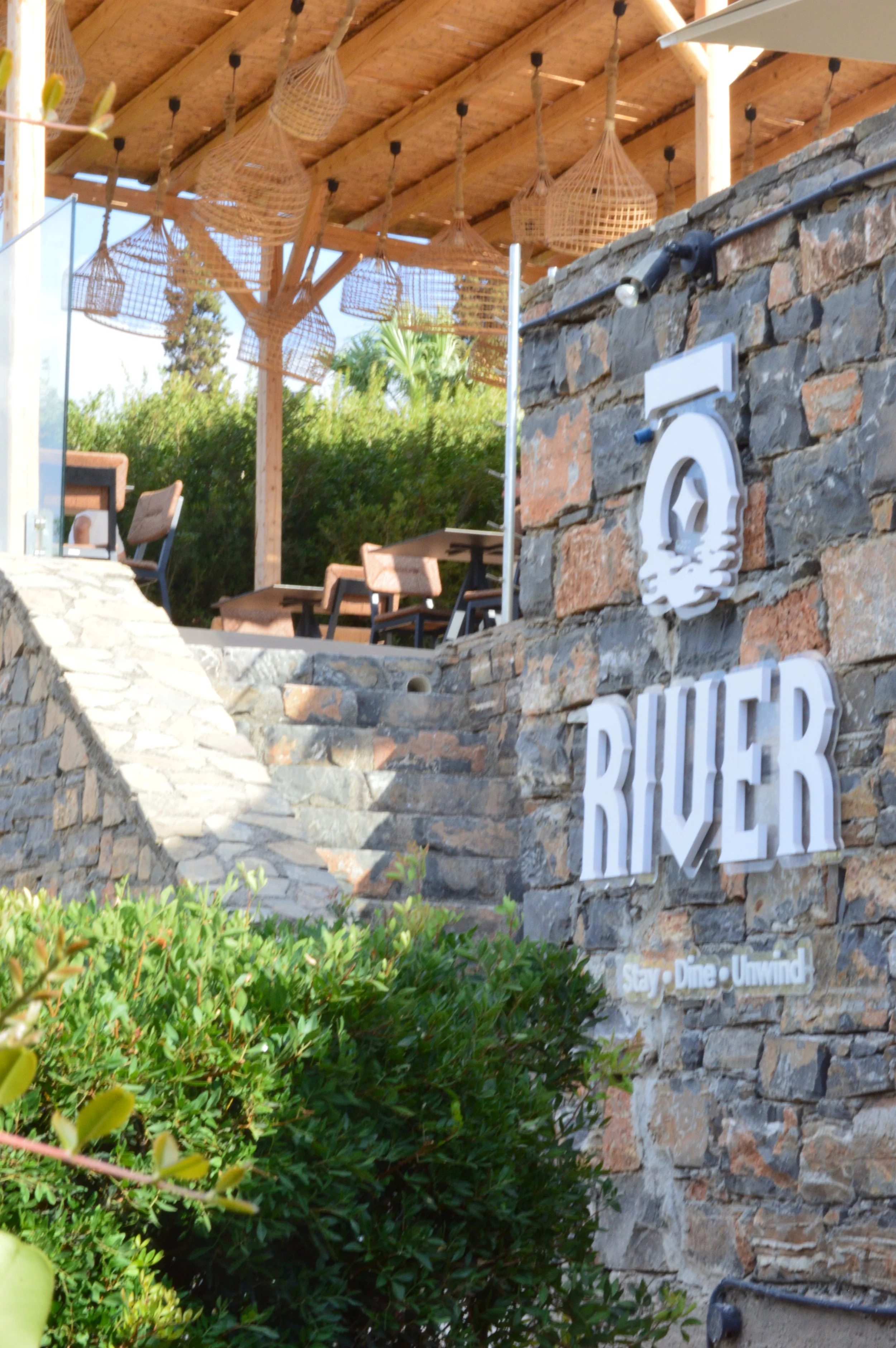 Exterior of a restaurant called River with outdoor seating, wooden tables and chairs, surrounded by greenery, and a stone wall with the restaurant's name and logo on it.