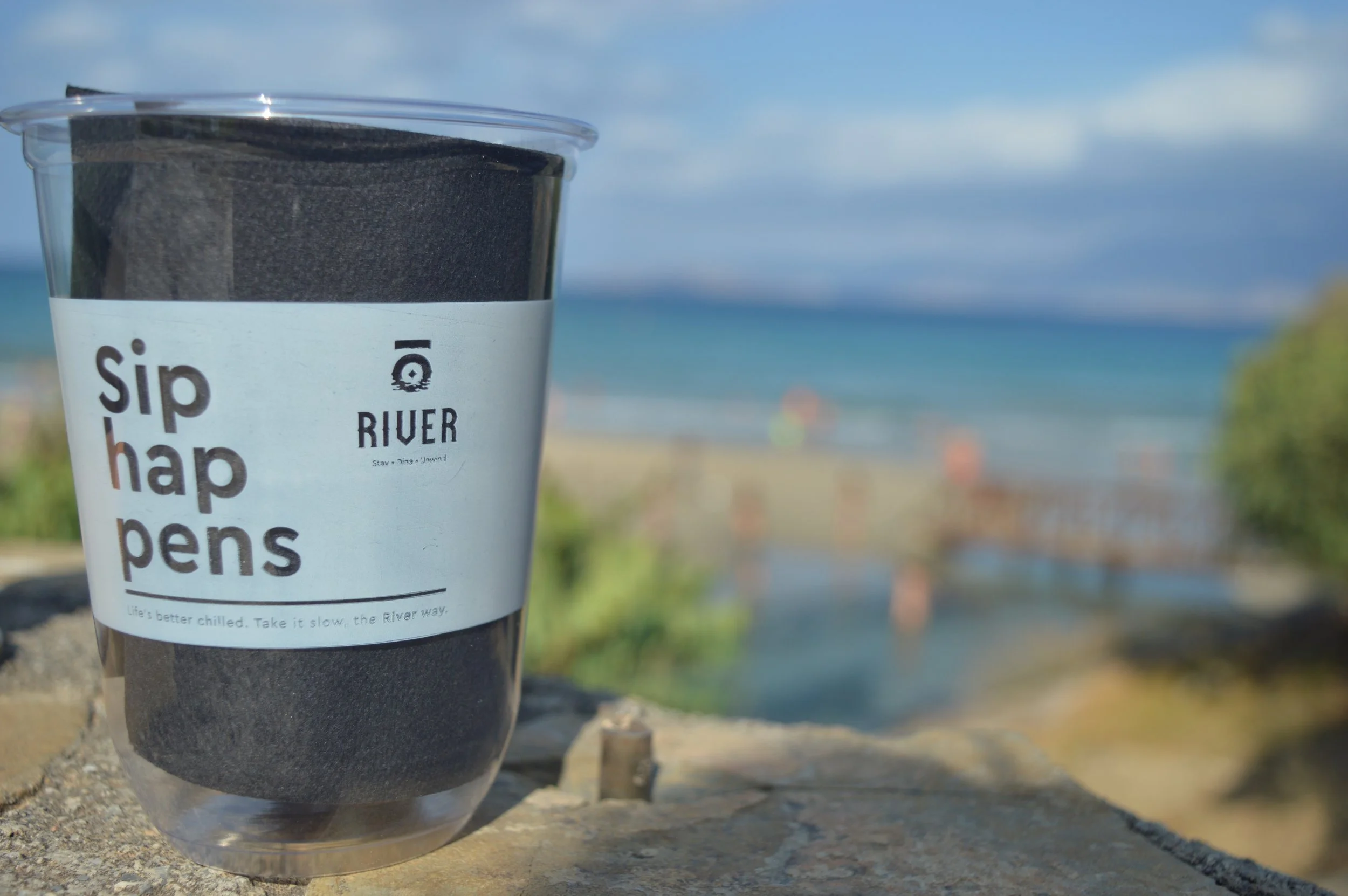 A clear plastic cup with a black filter inside, labeled 'sip hap pens' with a river scene in the background.