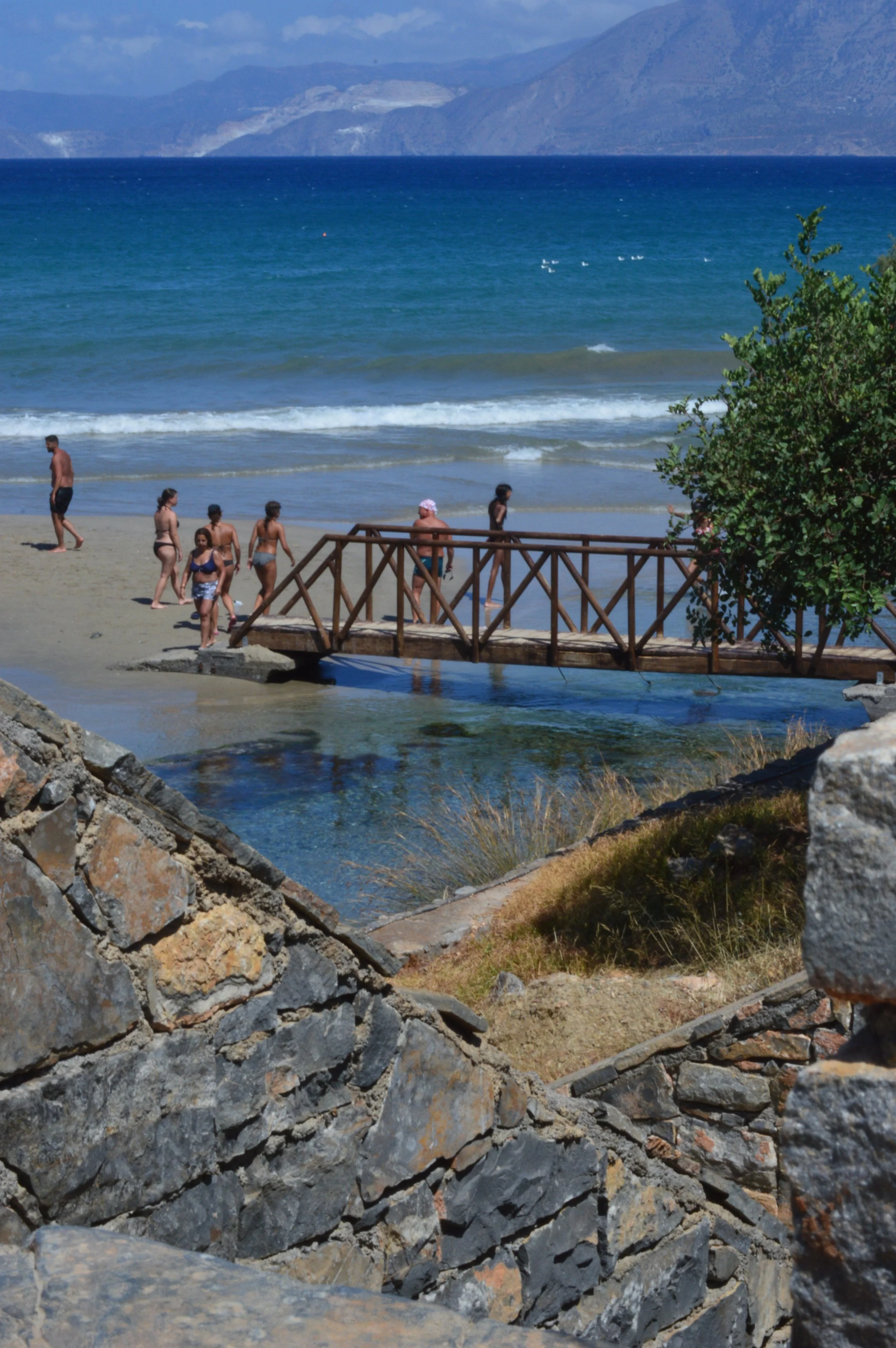 People on a sandy beach walking near a wooden bridge over a small stream, with ocean waves and distant mountains in the background.