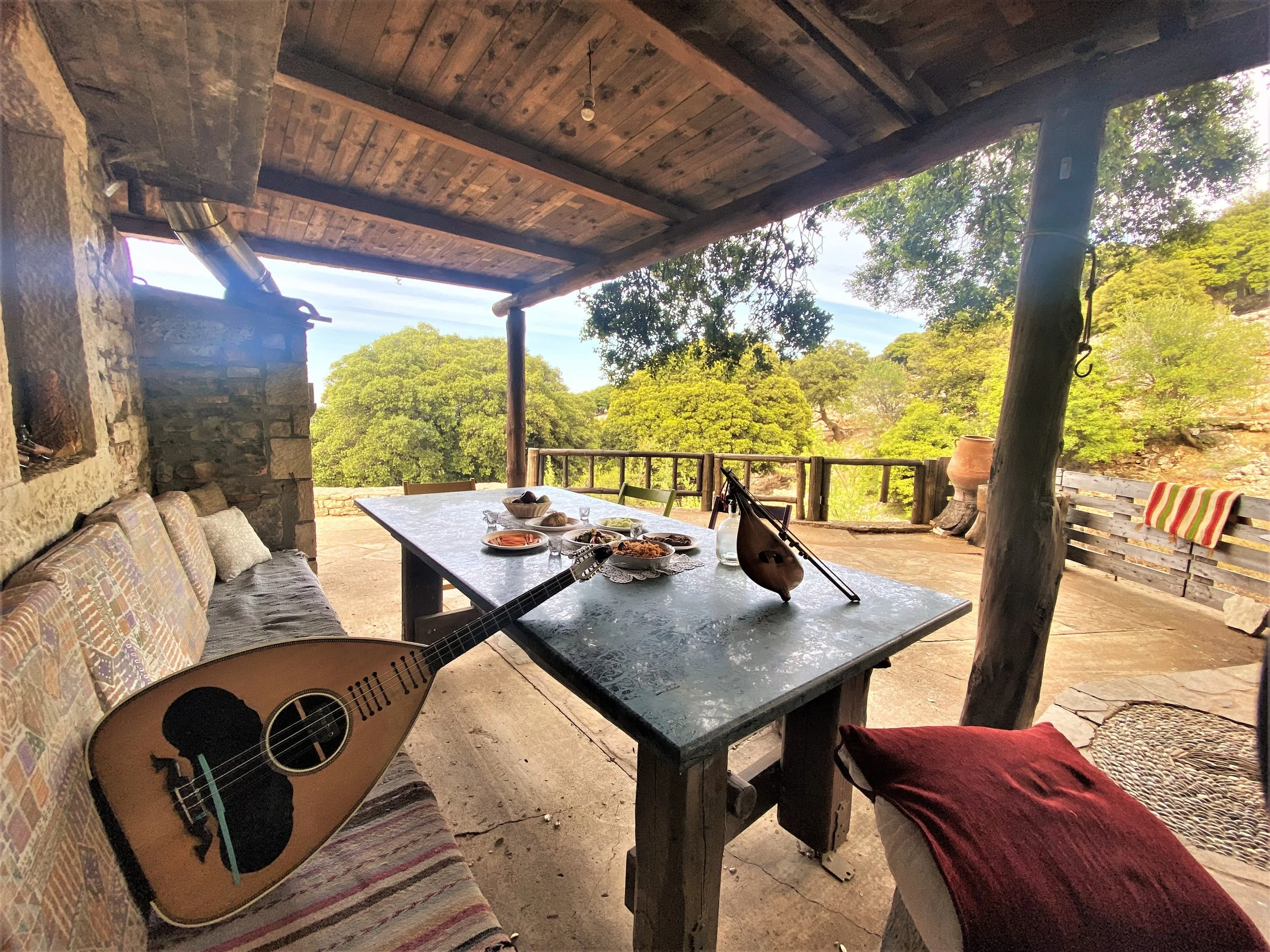 An outdoor rustic patio with a stone wall, wooden roof, and a table set with food and drinks. A musical instrument, a teardrop-shaped mandolin, is on the table, next to a smaller stringed instrument. There is a cushioned bench and a red cloth on a chair, with lush green trees in the background.