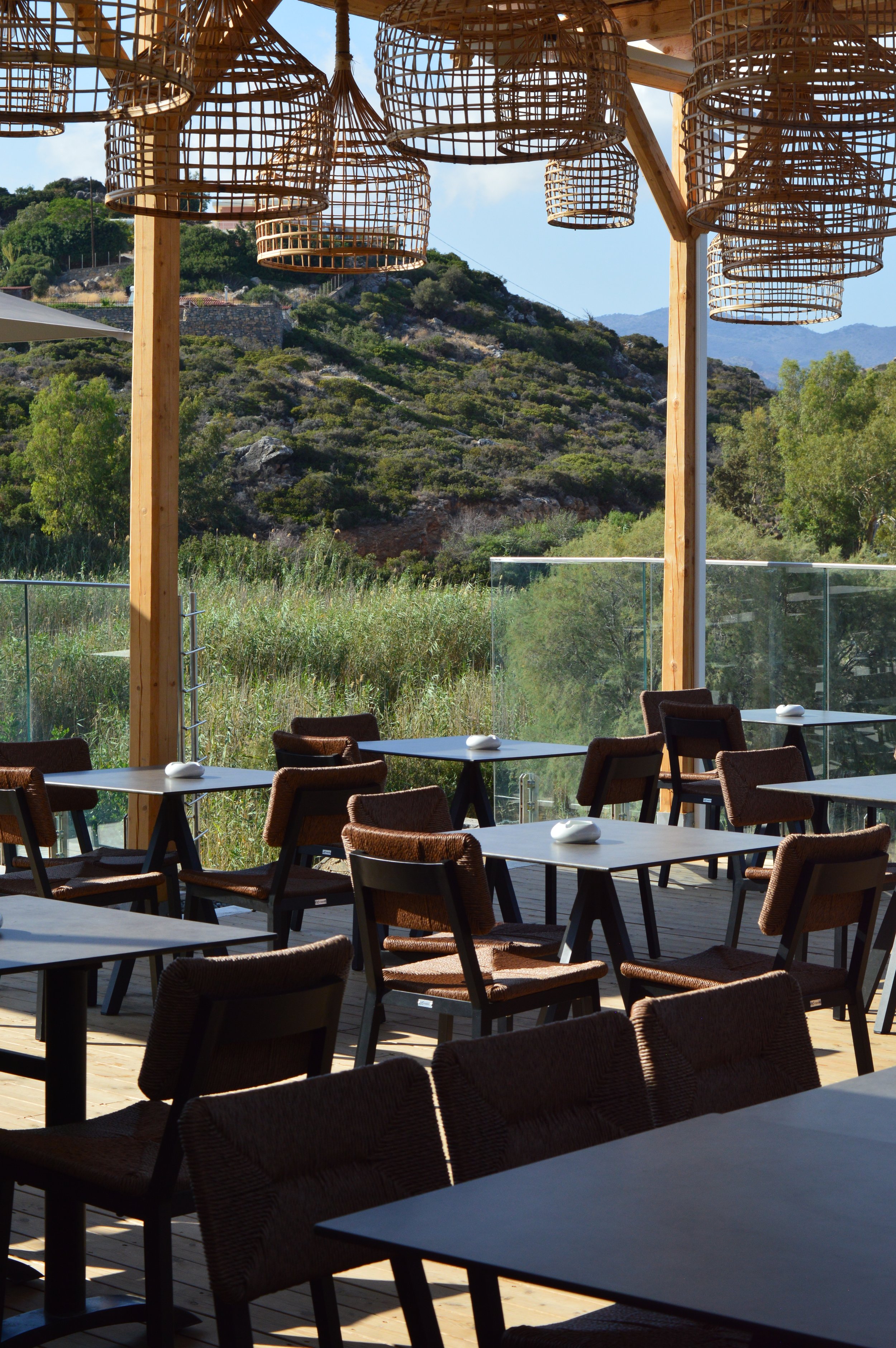 Outdoor restaurant with black tables and brown chairs, overlooking a lush green hillside with mountains in the background, under wicker hanging light fixtures.