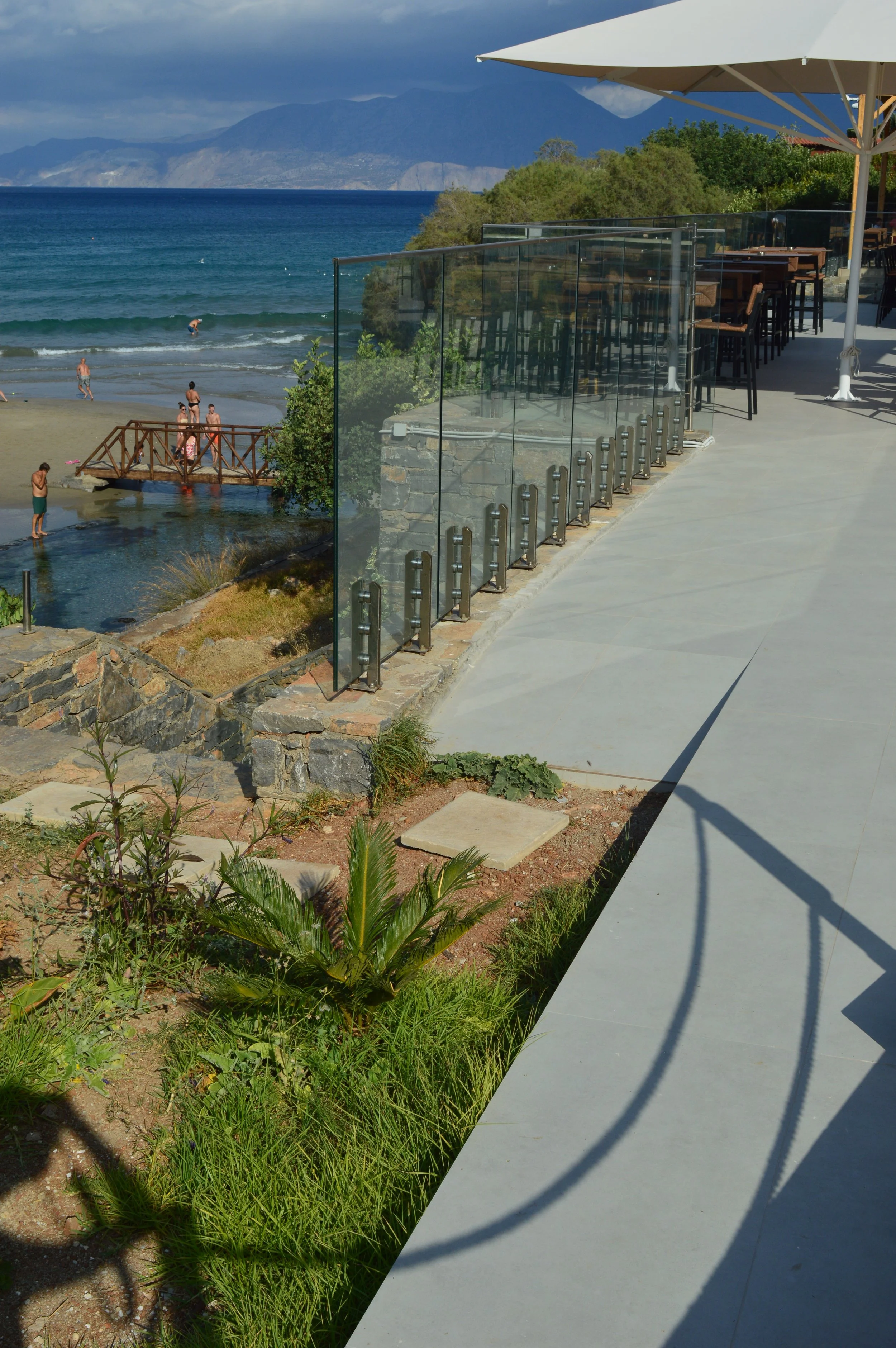 A beachfront patio with outdoor seating, glass railing, and an umbrella overlooking a beach with people swimming and a mountain in the background.