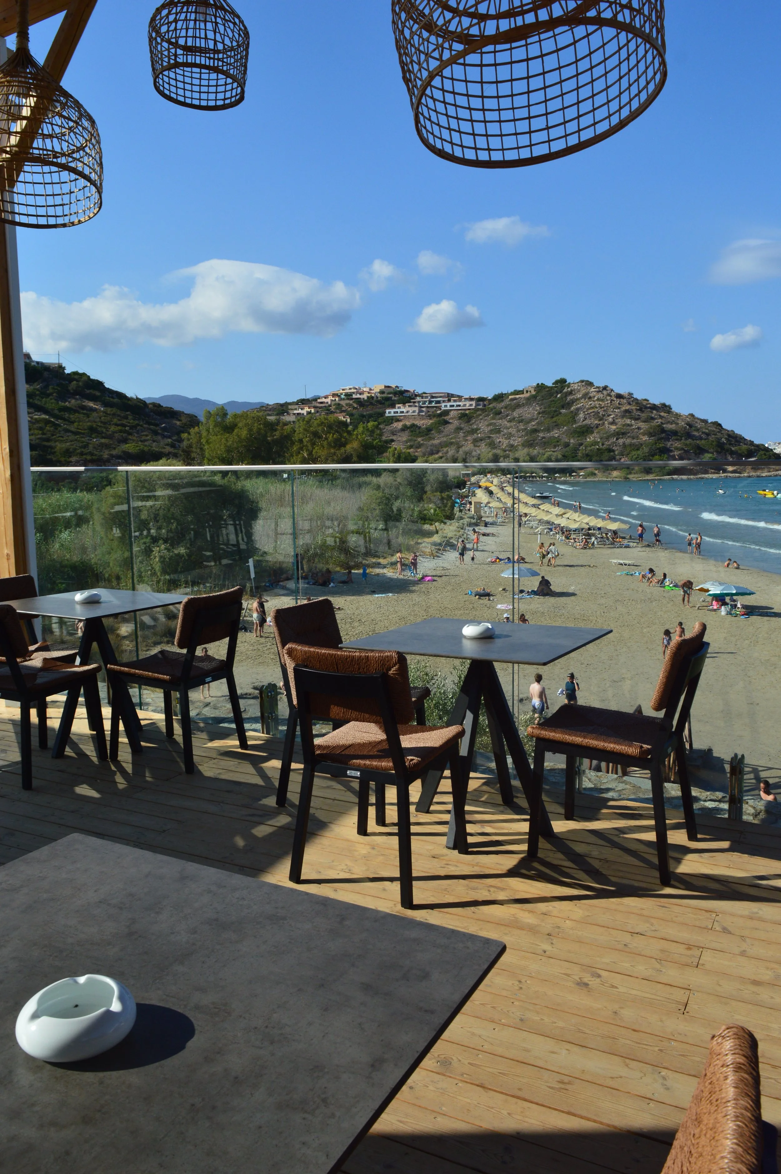 A view from inside a restaurant with a balcony overlooking a crowded beach with umbrellas, sunbathers, and boats in the water. There are wicker hanging lamps in the foreground.