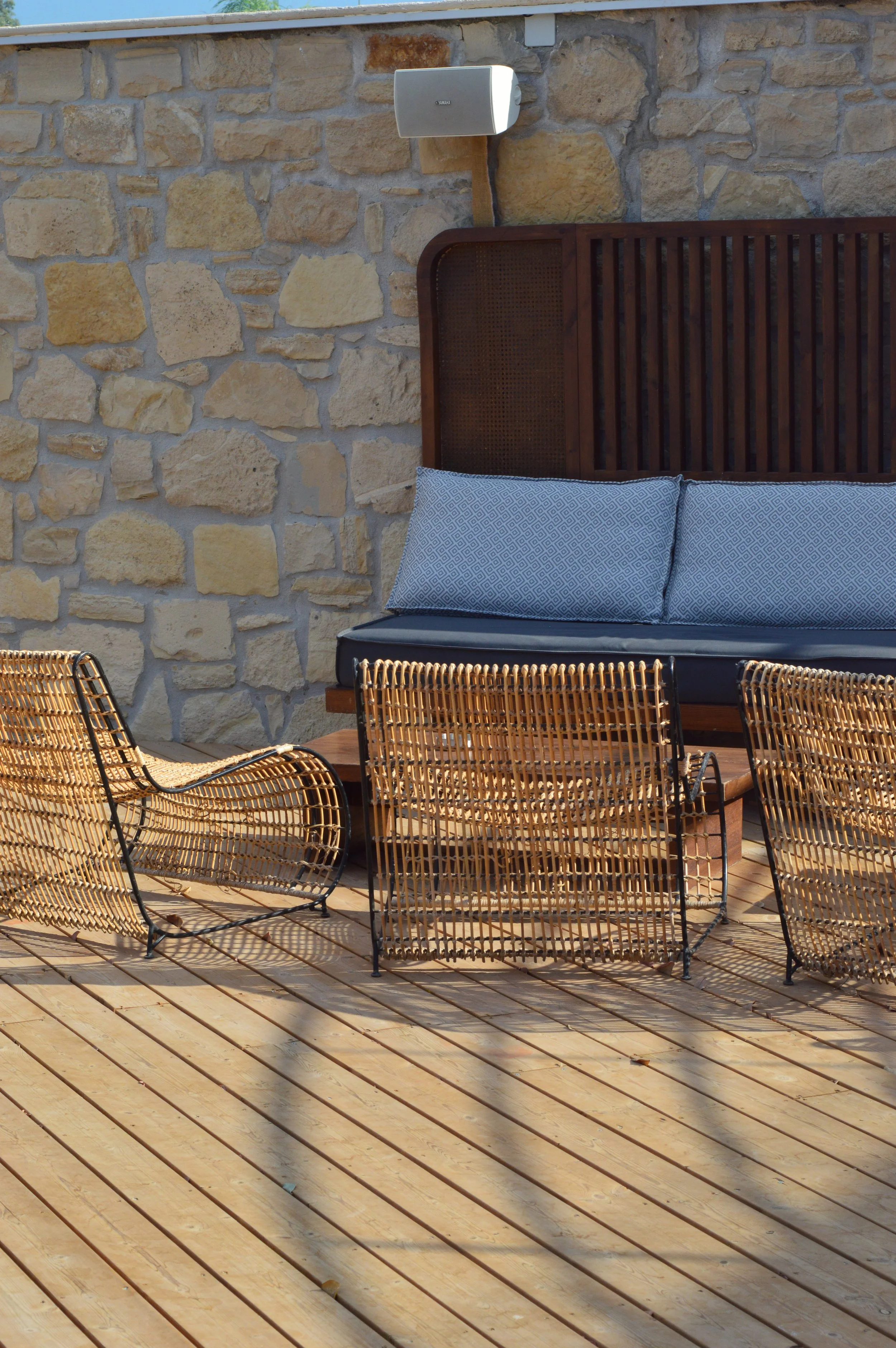 Outdoor patio area with rattan chairs, a wooden bench with cushions, a stone wall, and a speaker mounted on the wall.