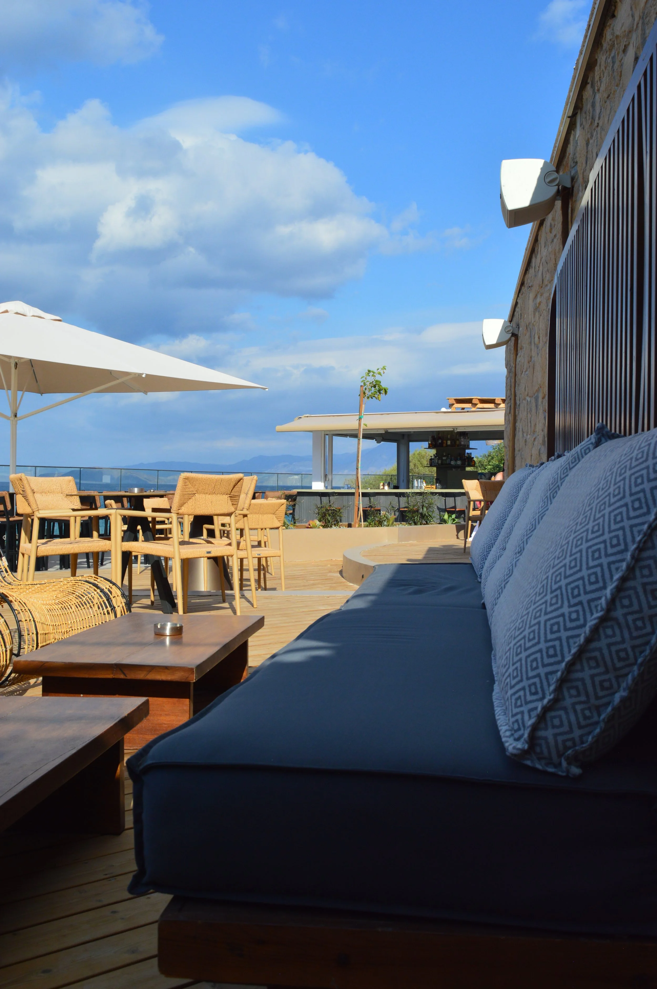 Outdoor rooftop patio with wicker chairs, tables, a large umbrella, a cushioned seating area, and a bar in the background under a blue sky with clouds.
