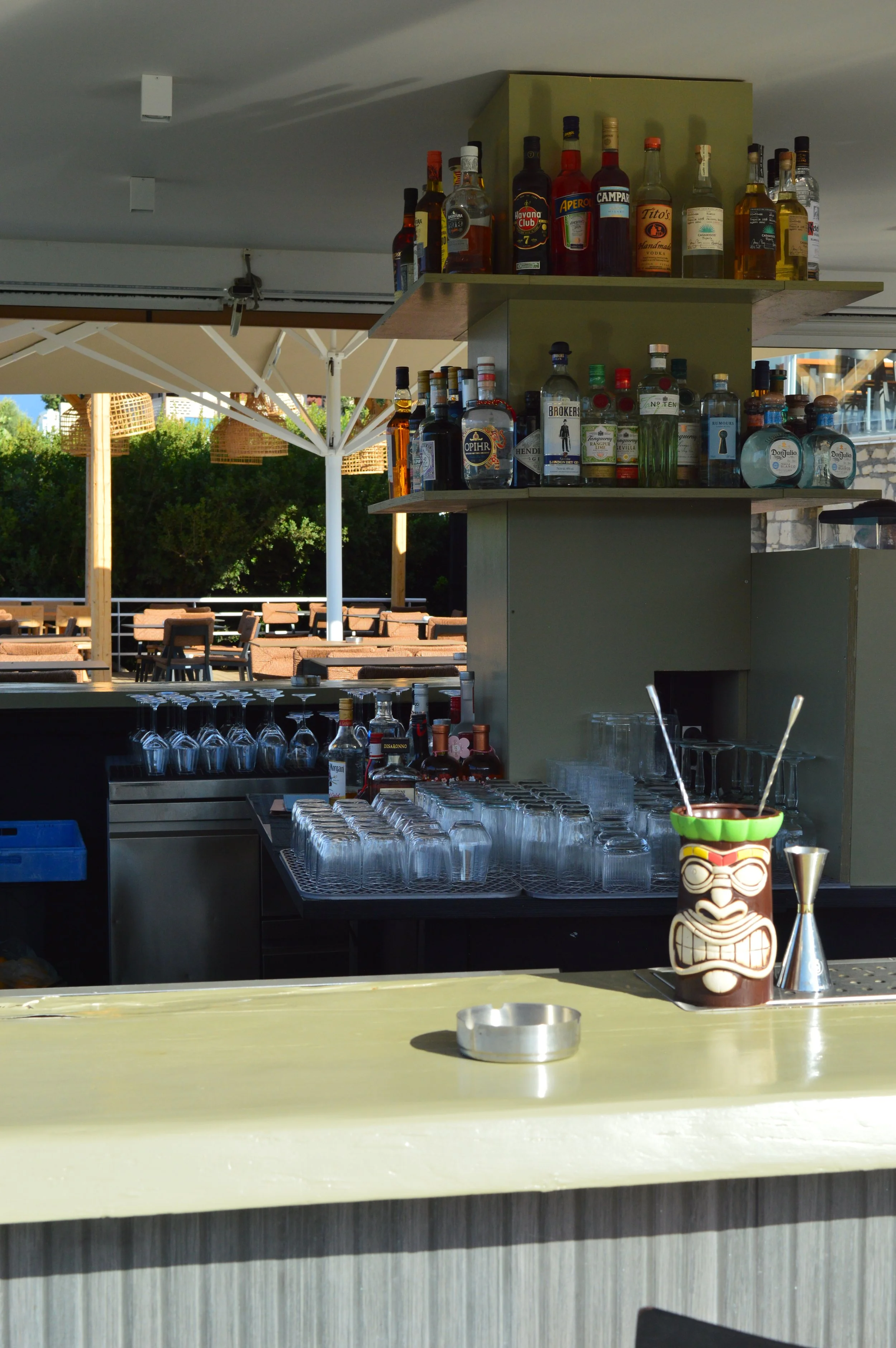 Bar area with liquor bottles on shelves, stacked glasses, a tiki mug, and an outdoor seating area with umbrellas in the background.