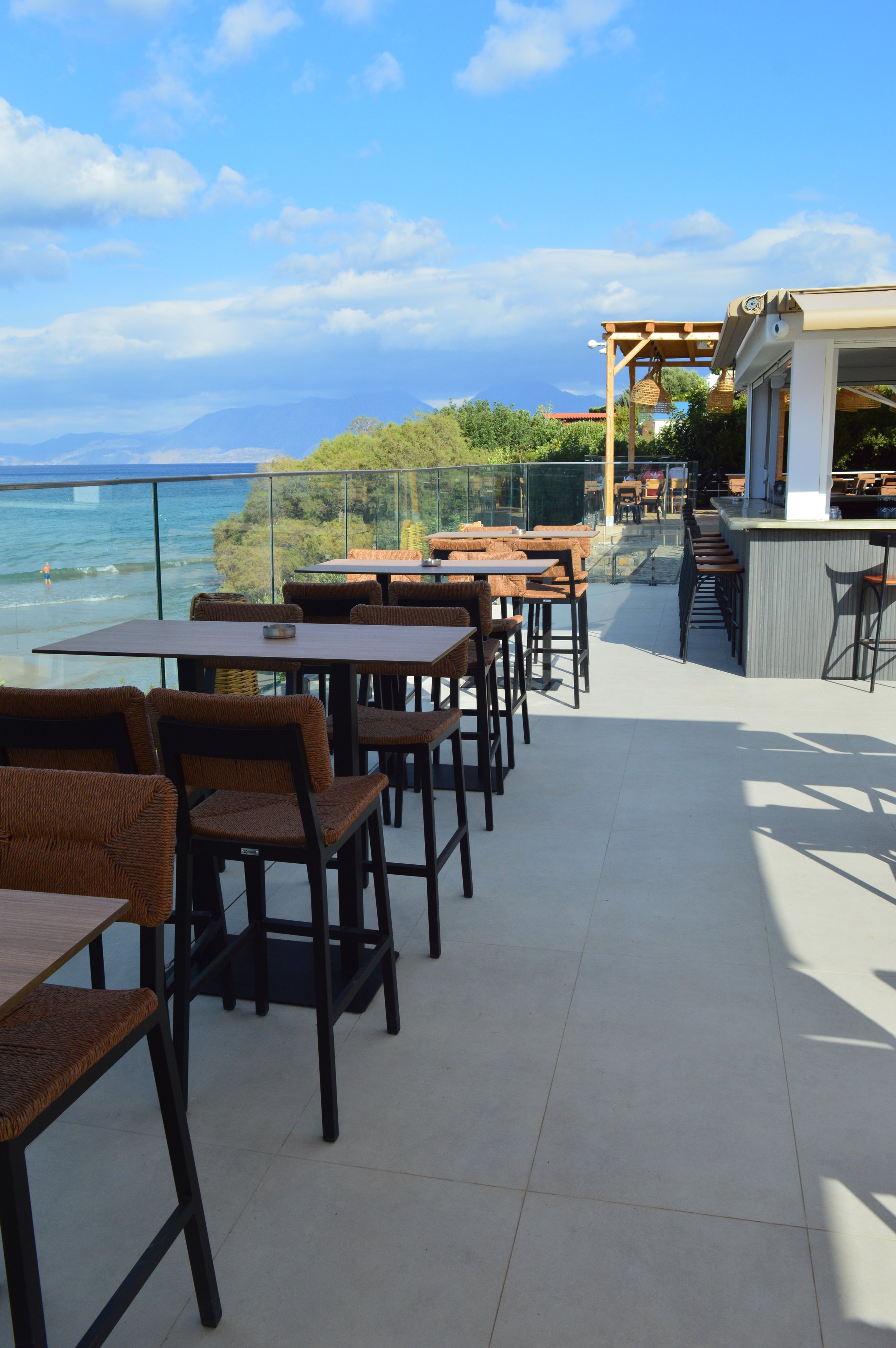Outdoor restaurant patio with wooden tables and chairs overlooking the ocean, with a glass barrier, partly shaded, and visible mountains and blue sky with clouds in the background.