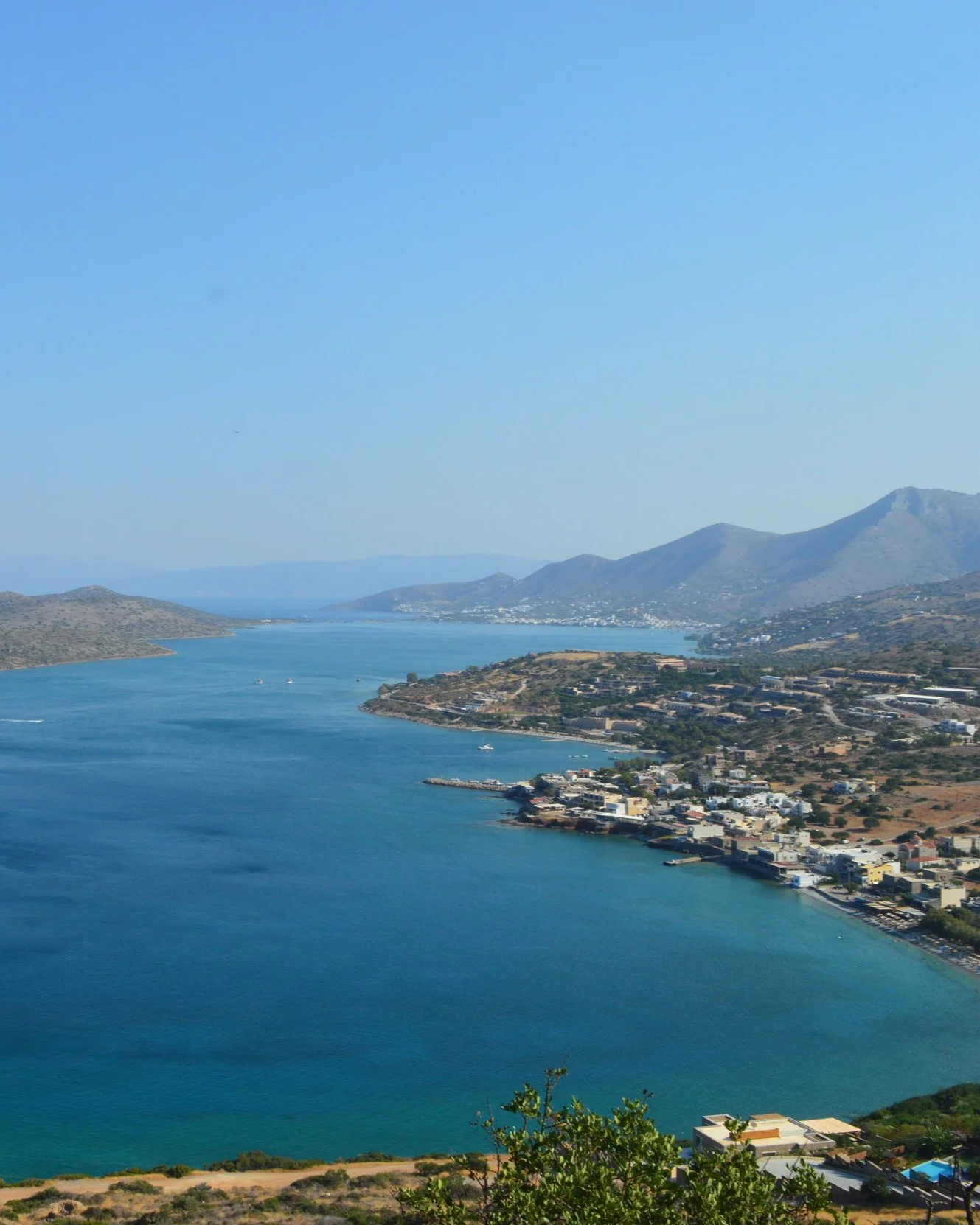 A scenic view of a large body of water, surrounded by hills and mountains, with a small town along the shoreline and boats on the water under a clear blue sky.