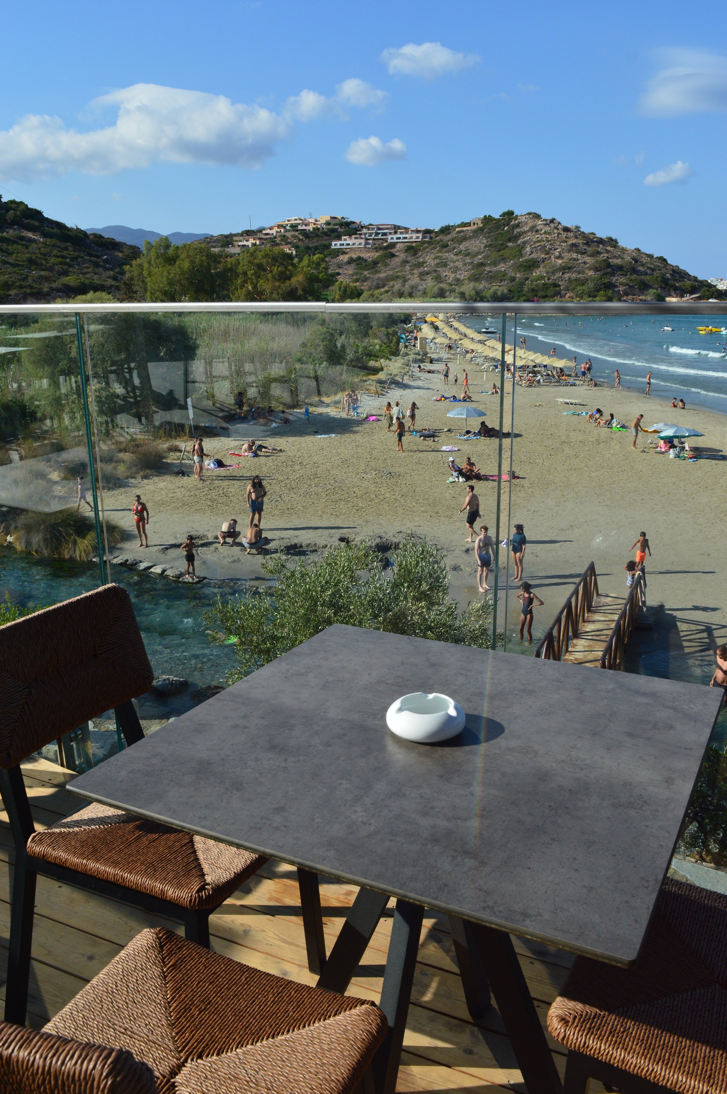 A beach scene viewed from a patio with a stone table and wicker chairs, a beach with umbrellas and people, and hills with buildings in the background.