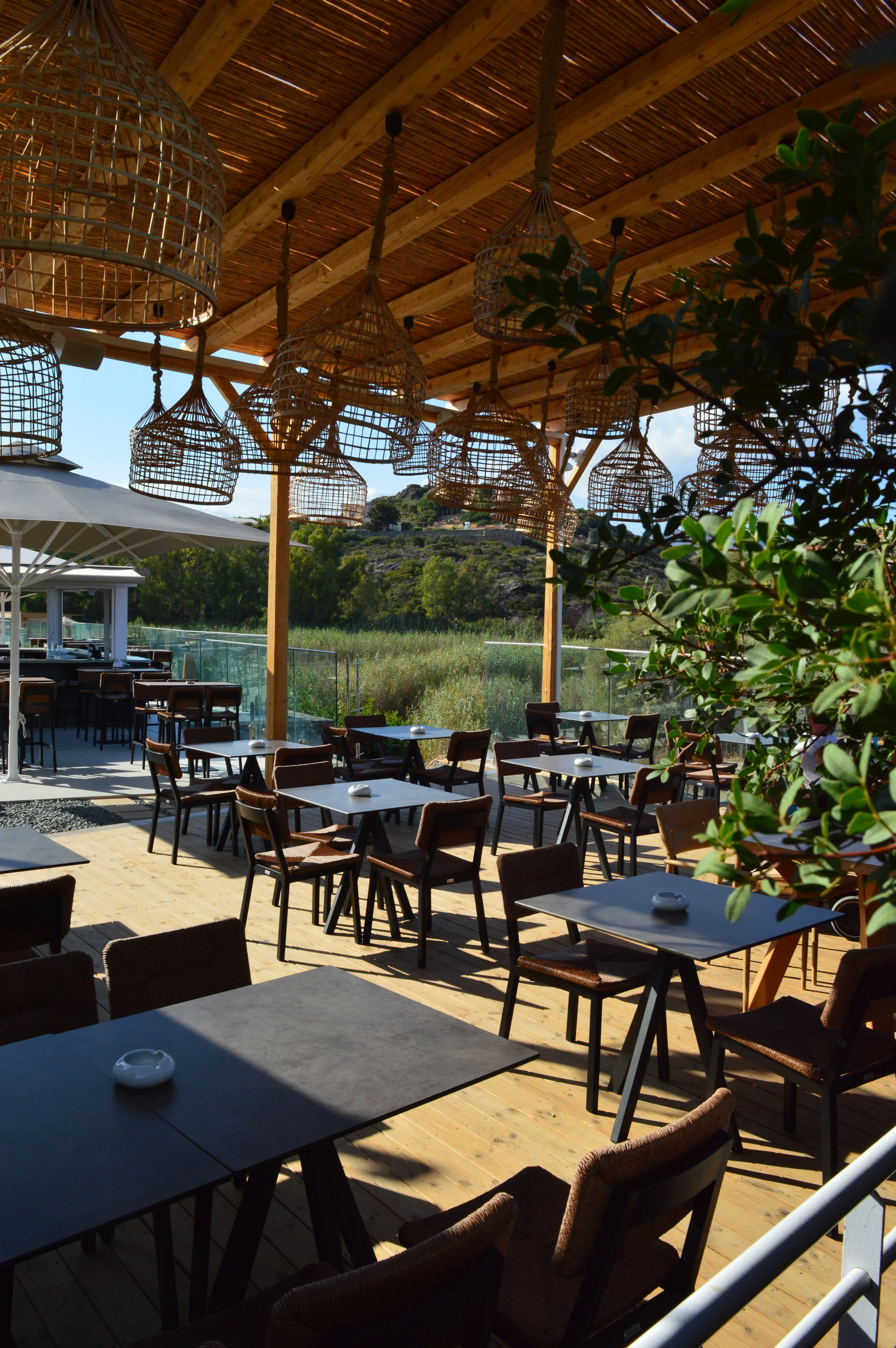 Outdoor dining area with black tables, brown chairs, and woven hanging light fixtures on a wooden deck, overlooking a green landscape and a partly cloudy sky.