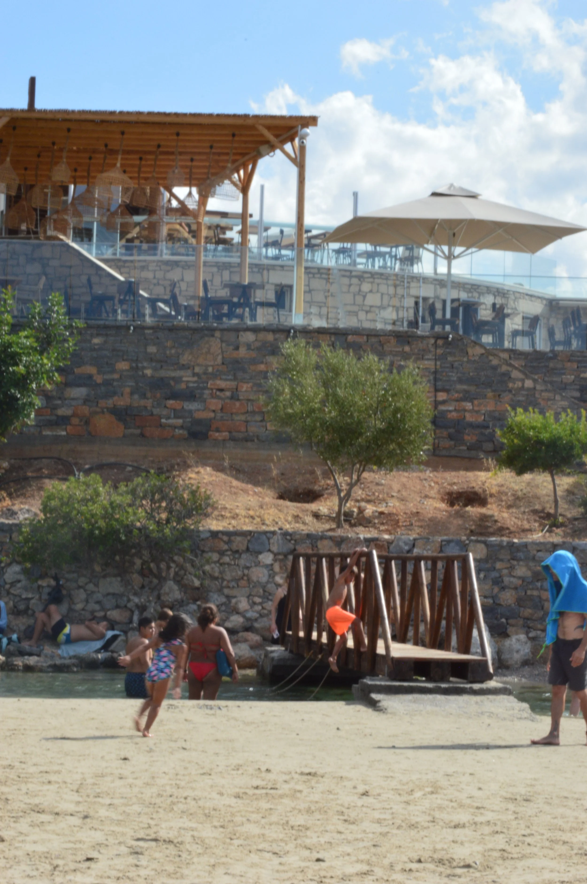 People playing and relaxing on a sandy beach near a small wooden dock with children climbing on it, and a rocky hillside with trees and an outdoor restaurant with umbrellas and tables on a sunny day.