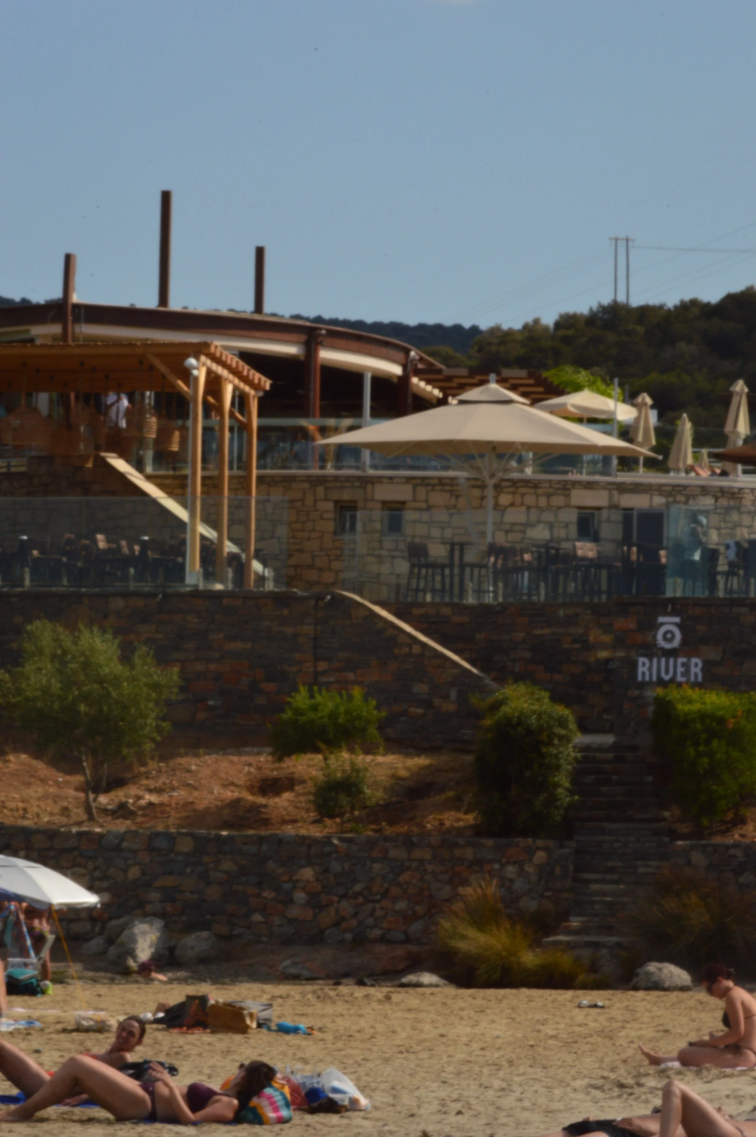 People lounging on the beach with a multi-tiered restaurant or bar on a hill in the background, with umbrellas and trees.