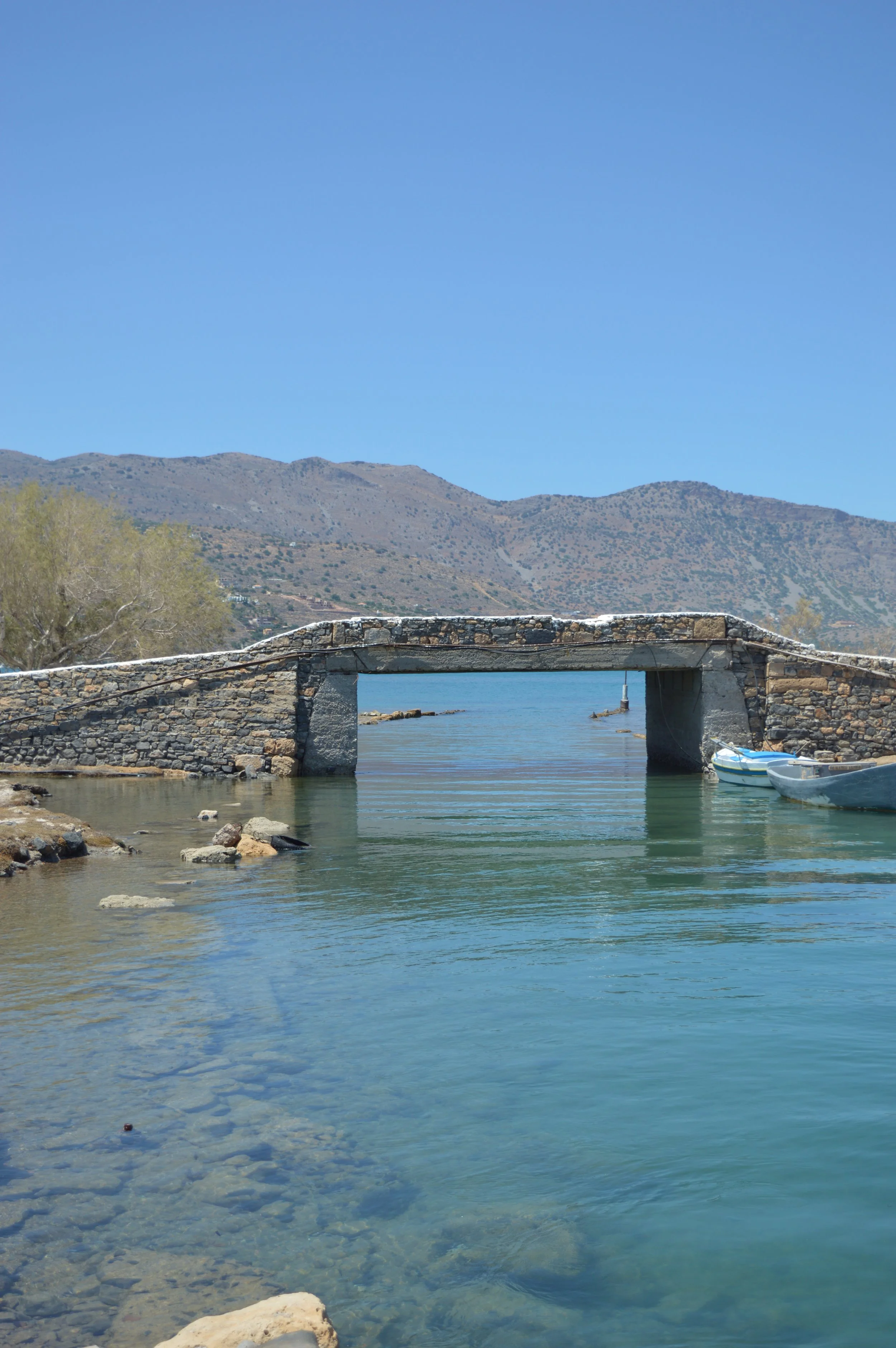 A stone bridge over calm water with small boats tied on the right side, hills in the background, and a clear blue sky.