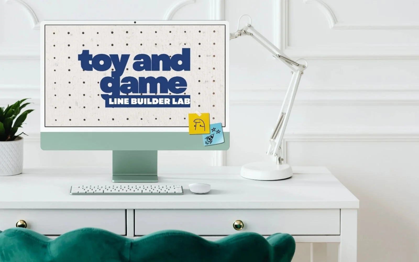 A computer monitor on a white desk displays the text 'toy and game line builder lab' with small sticky notes attached showing doodles of a face, stars, and a rocket. The desk also has a white keyboard, mouse, a white adjustable desk lamp, and a potted plant to the left.