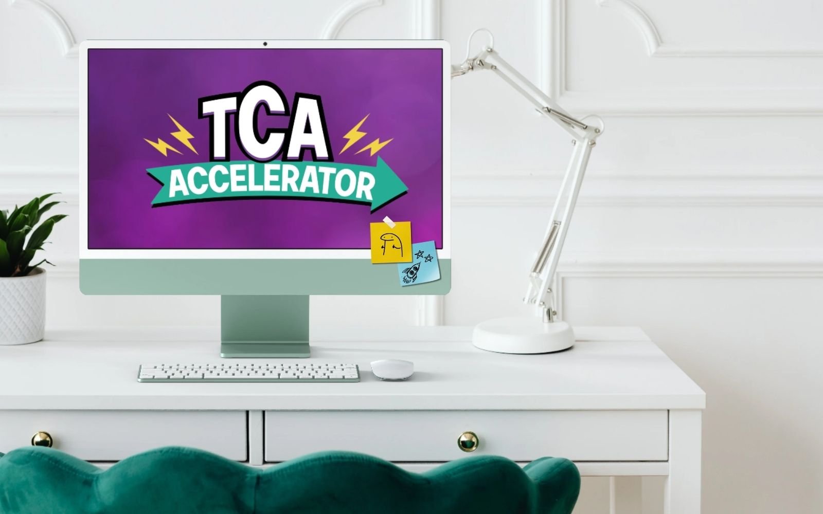 Computer monitor displaying the text 'TCA Accelerator' with a purple background, notes attached to the monitor, a white keyboard, a white mouse, a white desk with green drawers, a white desk lamp, and a potted plant on a white desk in a bright room.
