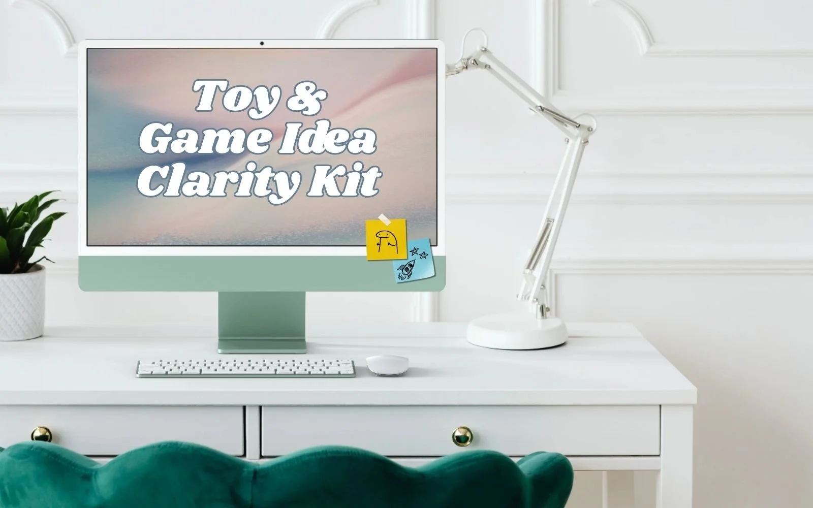 A computer monitor on a white desk displaying the text "Toy & Game Idea Clarity Kit" with colorful sticky notes attached to the bottom right corner, a white keyboard and mouse in front, a white adjustable desk lamp to the right, and a potted green plant on the left, against a white wall background.