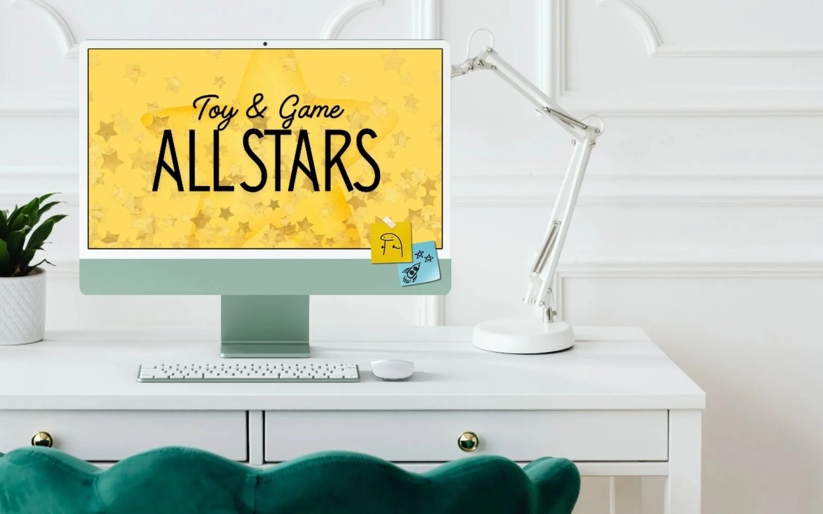 Computer monitor displaying a yellow background with star patterns and text that reads "Toy & Game All Stars" on a white desk with a white keyboard, mouse, white desk lamp, and a potted plant, in a bright room with white walls.