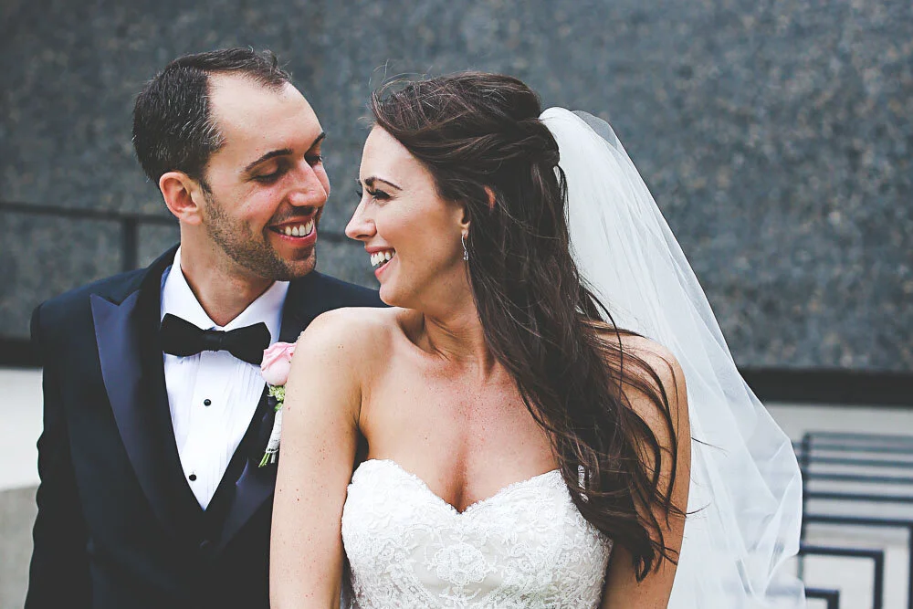 A bride and groom in wedding attire look at each other smiling.