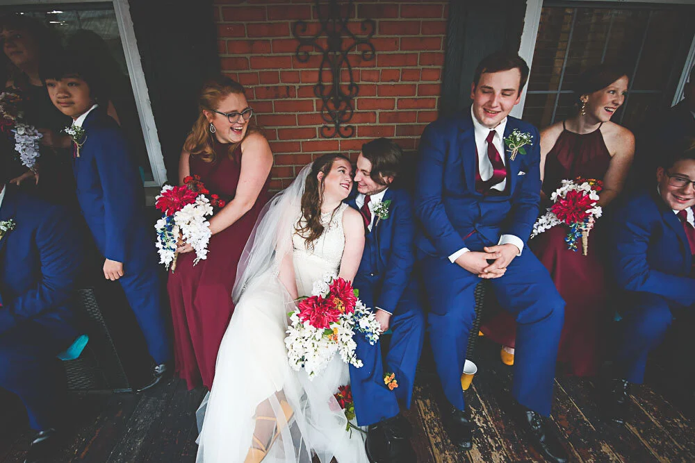 A bride and groom sit with faces close together on a bench in between their wedding party.