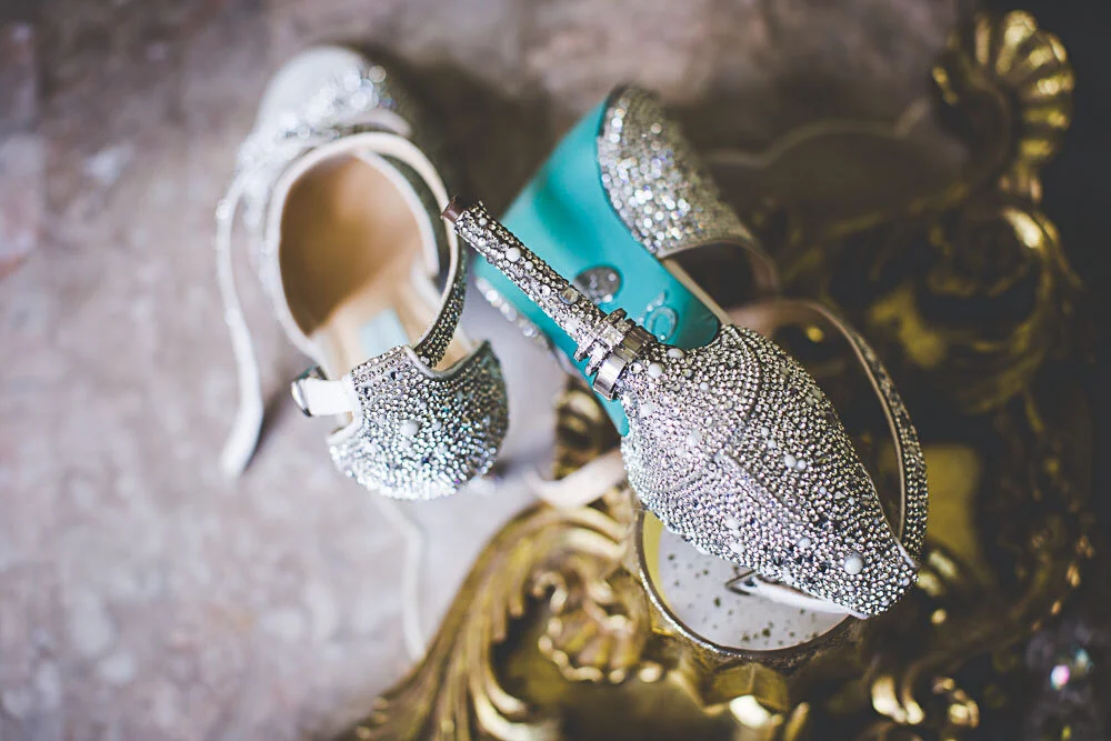 Wedding detail shot of rhinestone-covered bridal shoes with the wedding rings threaded on to the stiletto. An ornate gold table is in the background.