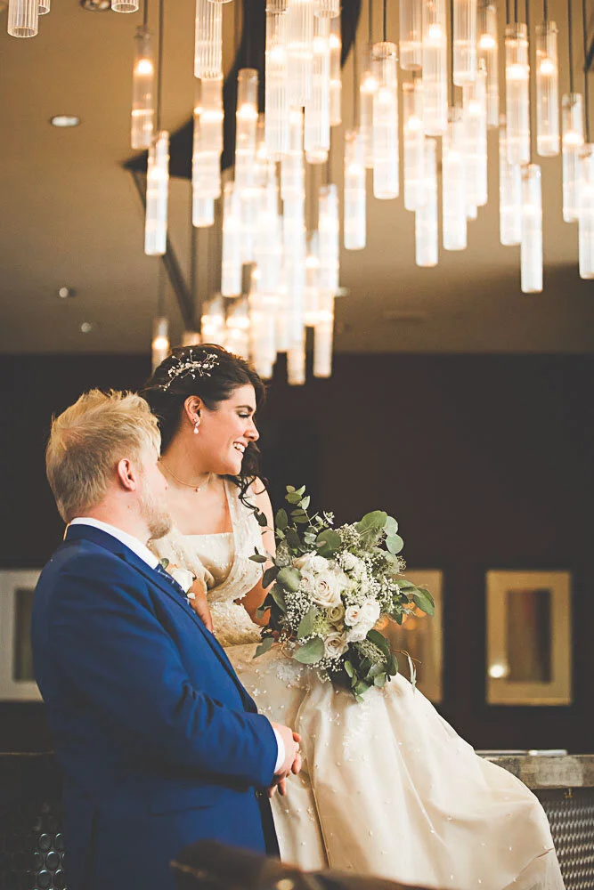 A bride and groom sit romantically in profile-view with a beautiful light fixture that looks like many candles hanging from the ceiling.