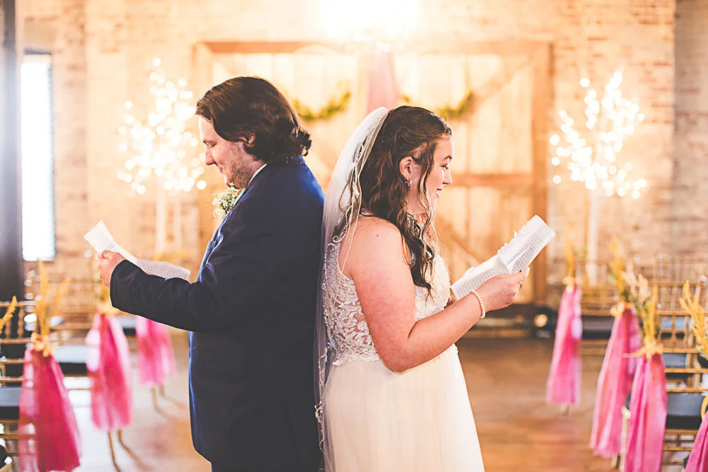 A bride and groom stand back to back while reading letters they have written to each other in their wedding venue before their ceremony.
