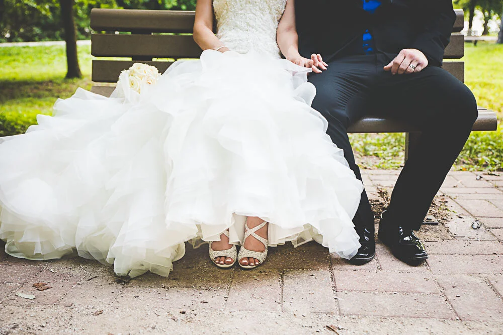 A bride and groom sit on a park bench side by side from the chest down, focusing on their attire and shoes.