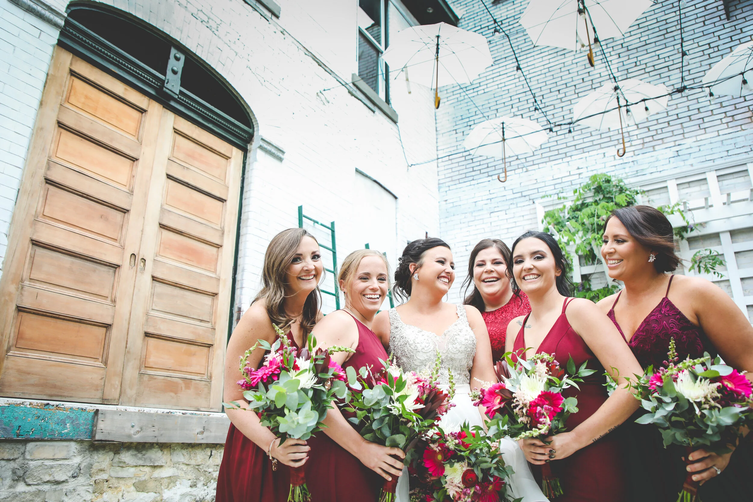 A bridal party stands close together and laughs under an open ceiling at Majorette with many white umbrellas hanging from the rooftop.